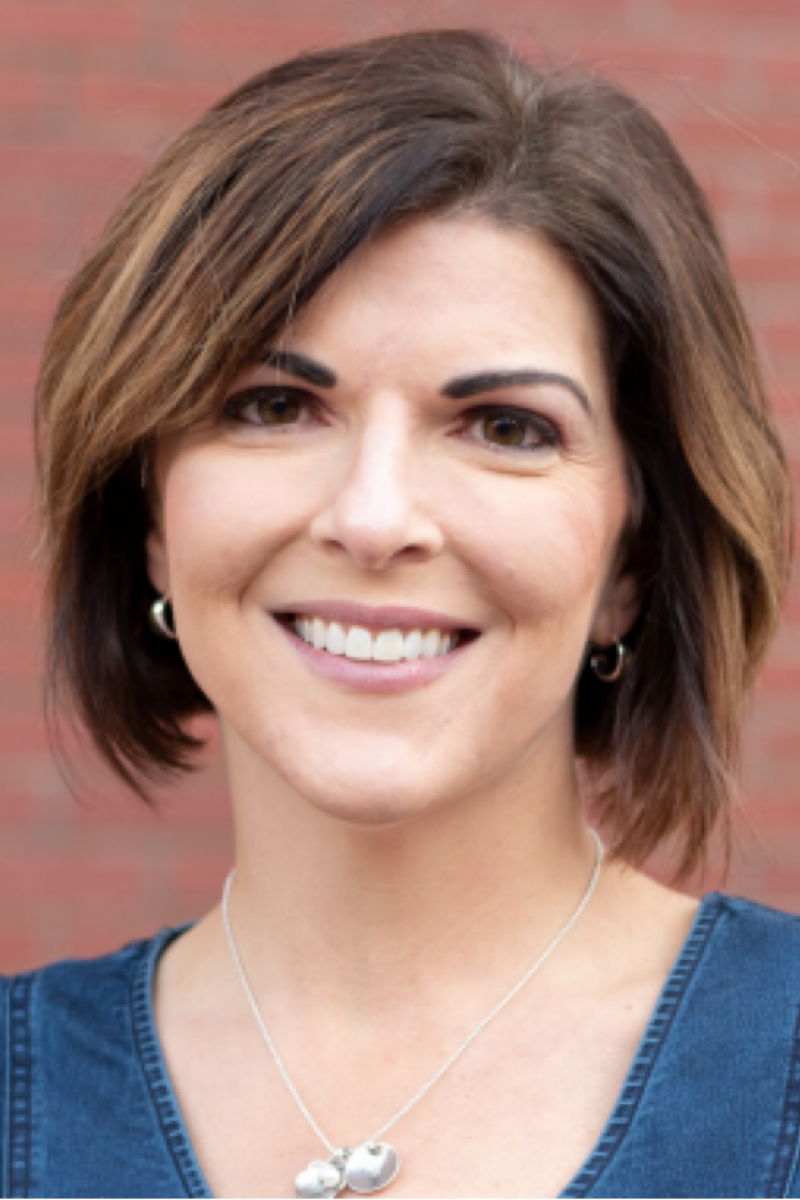 A woman wearing a necklace and earrings is smiling in front of a brick wall.