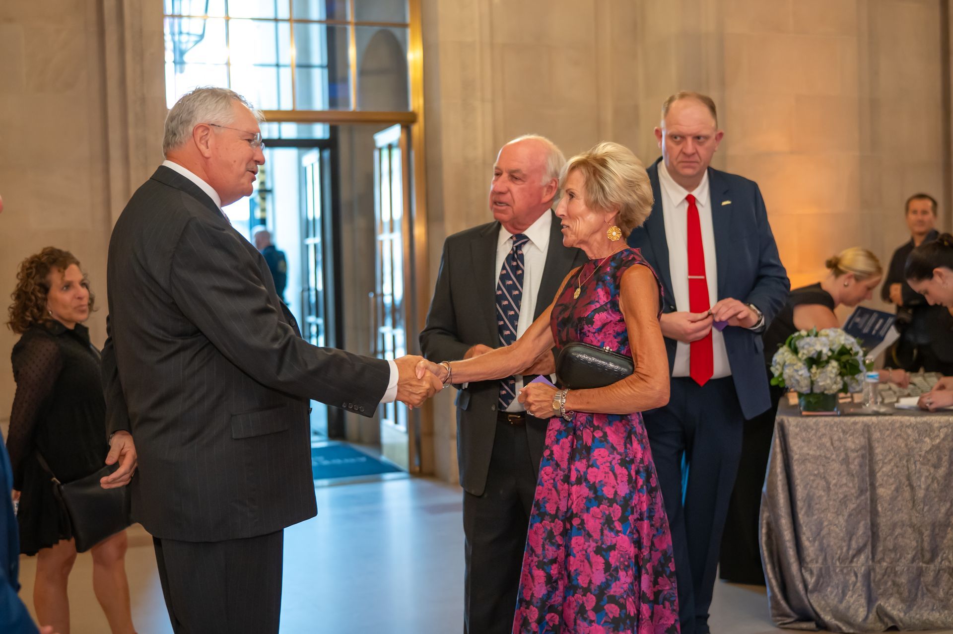 Two men shake hands at a formal indoor event, with guests standing nearby.