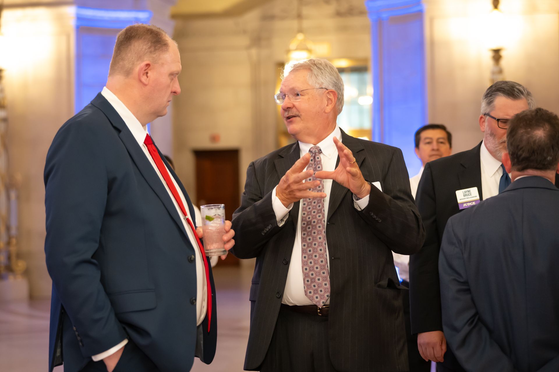 Two men in suits chatting at a formal indoor event, one holding a drink, with others in the background