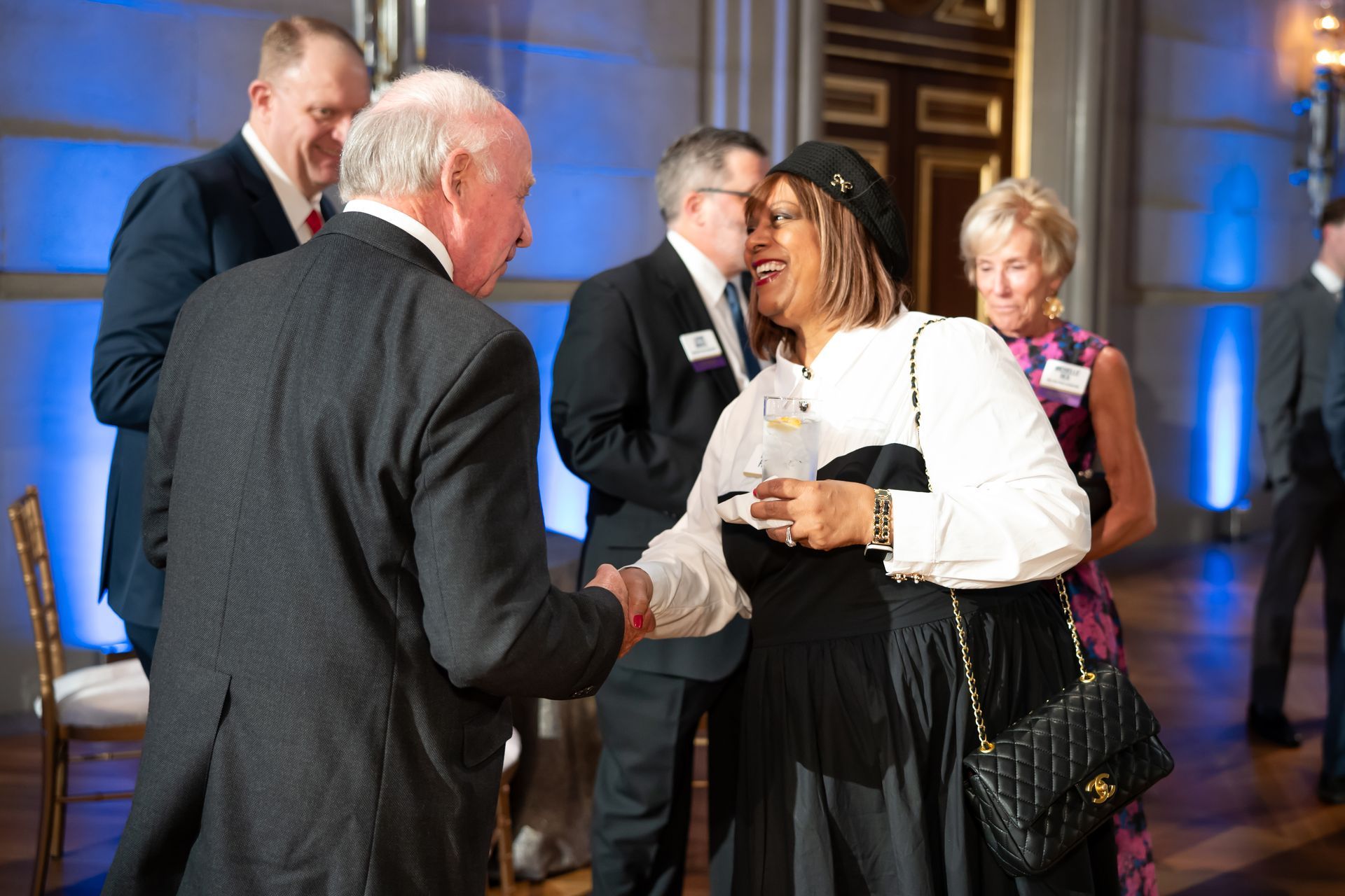 Guests in formal attire chat at a reception in a warmly lit ballroom.