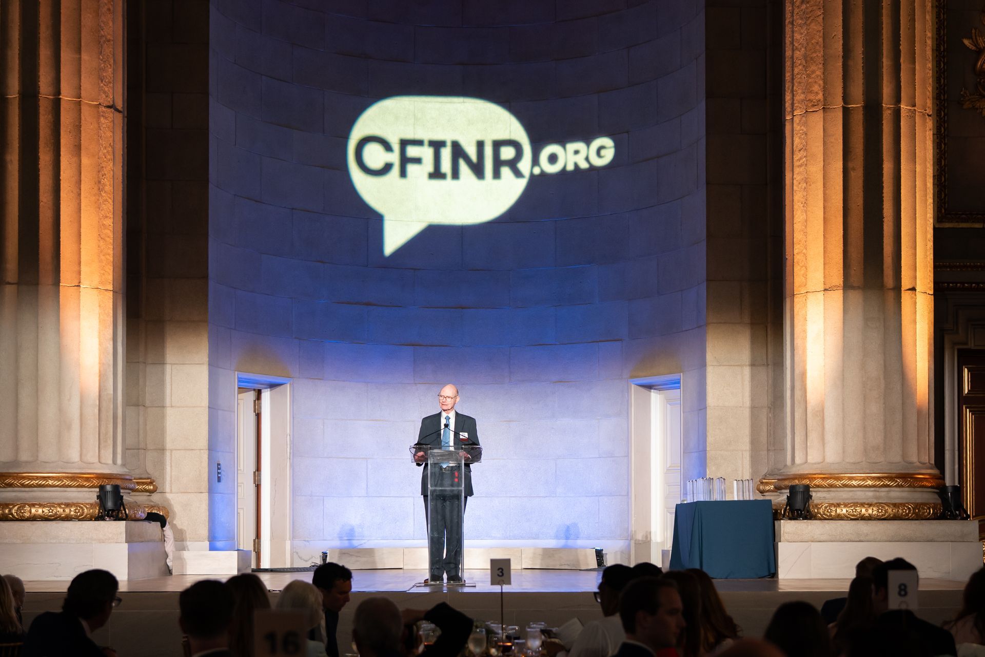 Speaker on stage beneath large CFINR.org banner in a dim auditorium