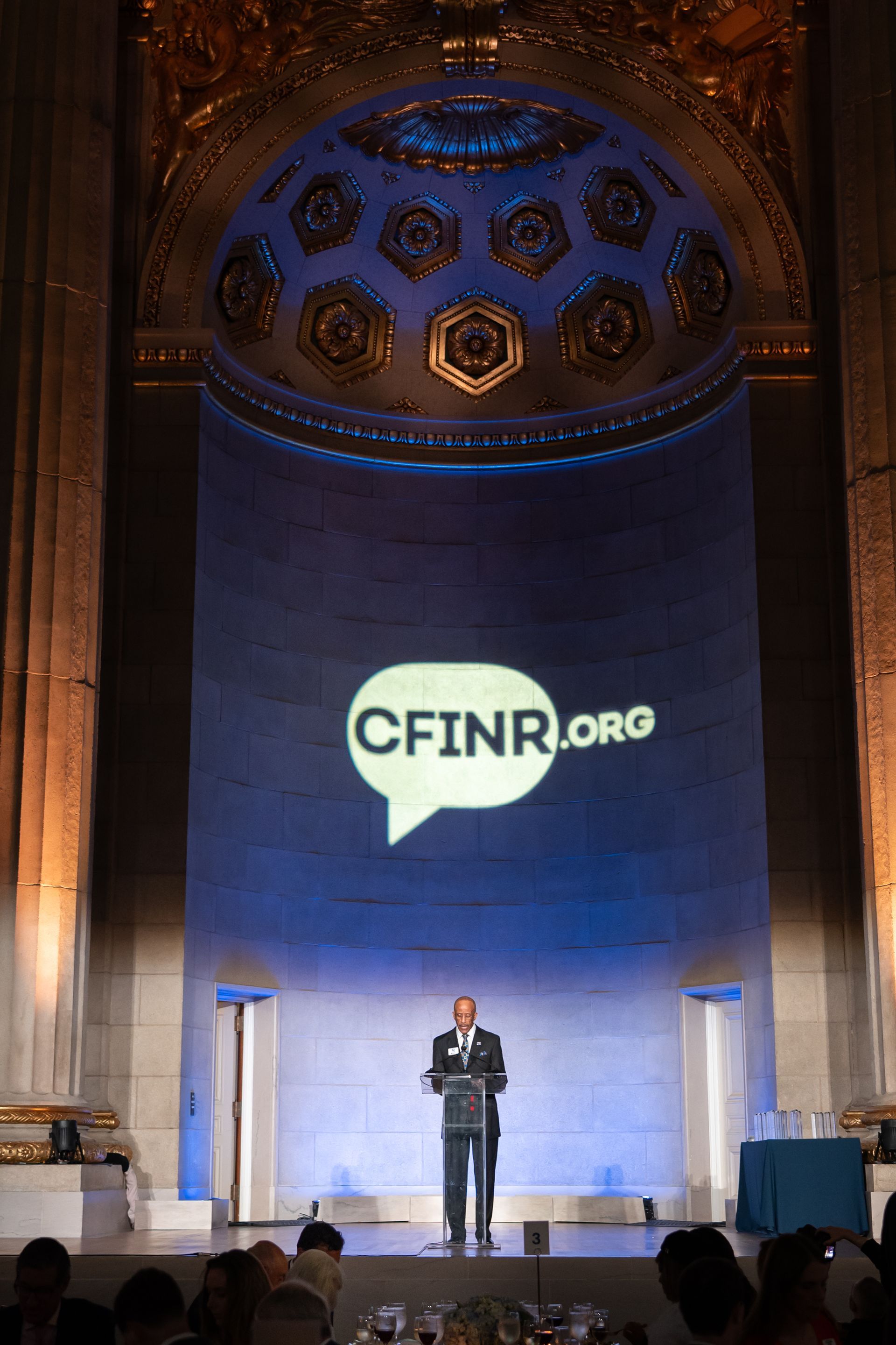 Speaker on stage beneath large CFINR.org projection in an ornate hall
