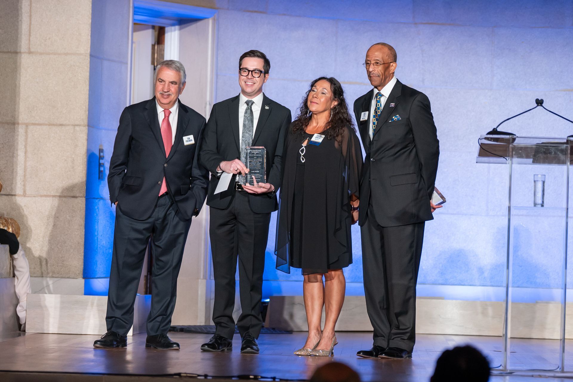 Four people stand on a stage holding an award, dressed in formal attire, with a podium and blue lighting behind them.