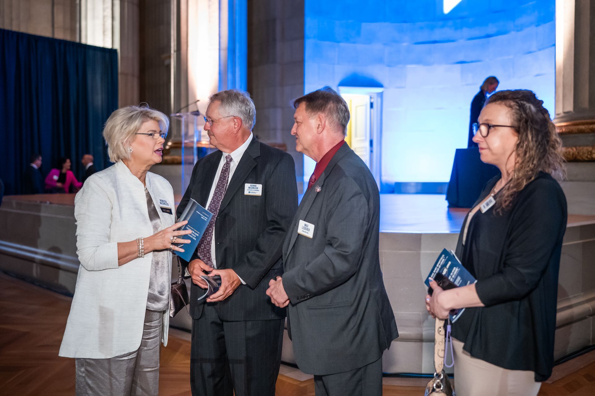 Four adults in business attire converse at a conference reception with a blue-lit backdrop.