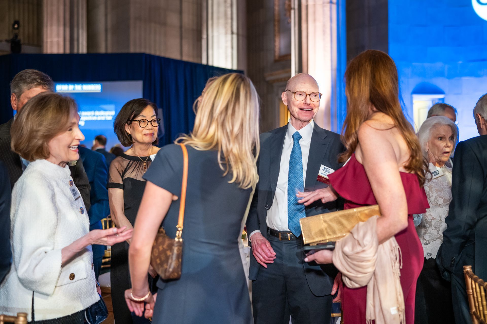 People mingling at a formal indoor event, with one woman holding a bouquet and others conversing nearby.