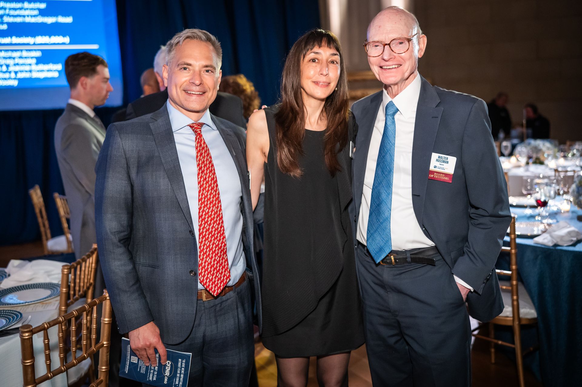 Three people smiling at a formal banquet, standing beside round tables with blue tablecloths