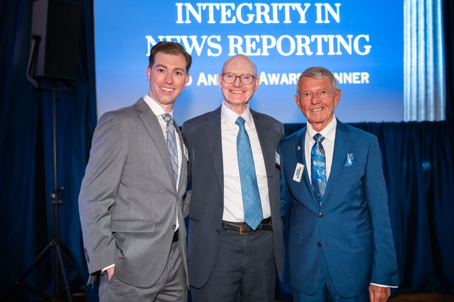 Three men in suits pose on stage before an “Integrity in News Reporting” award backdrop.