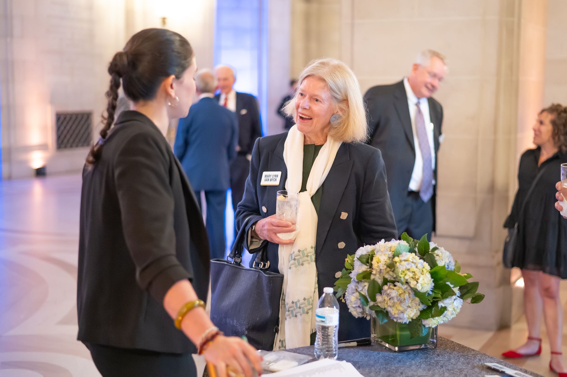 Two women chatting at a formal indoor event beside a table with flowers.