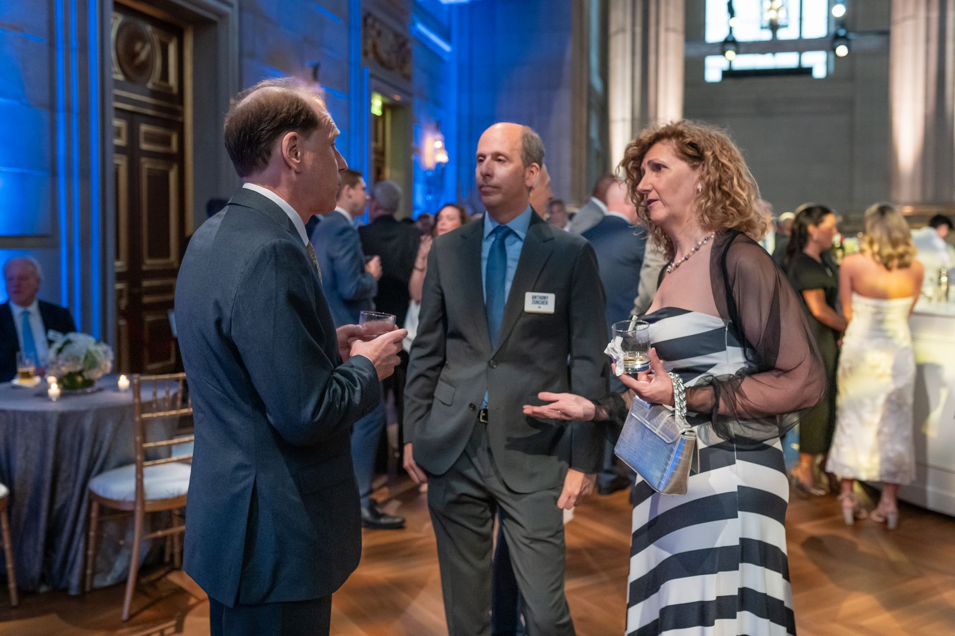 Three people in formal wear converse at a reception in a grand hall.