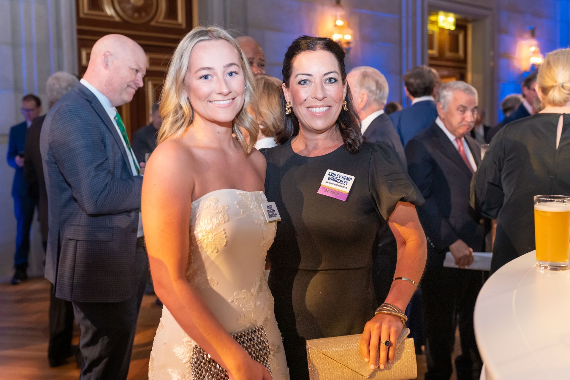 Two women posing at a formal event, with drinks and guests in the background