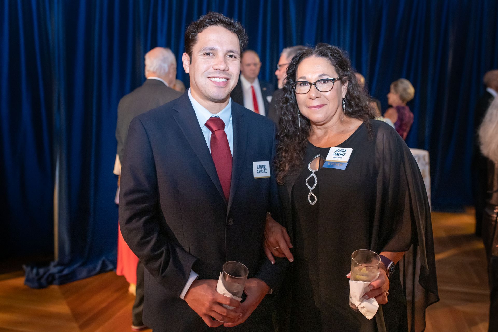 Two people in formal attire holding awards at an indoor event, standing in front of blue curtains.