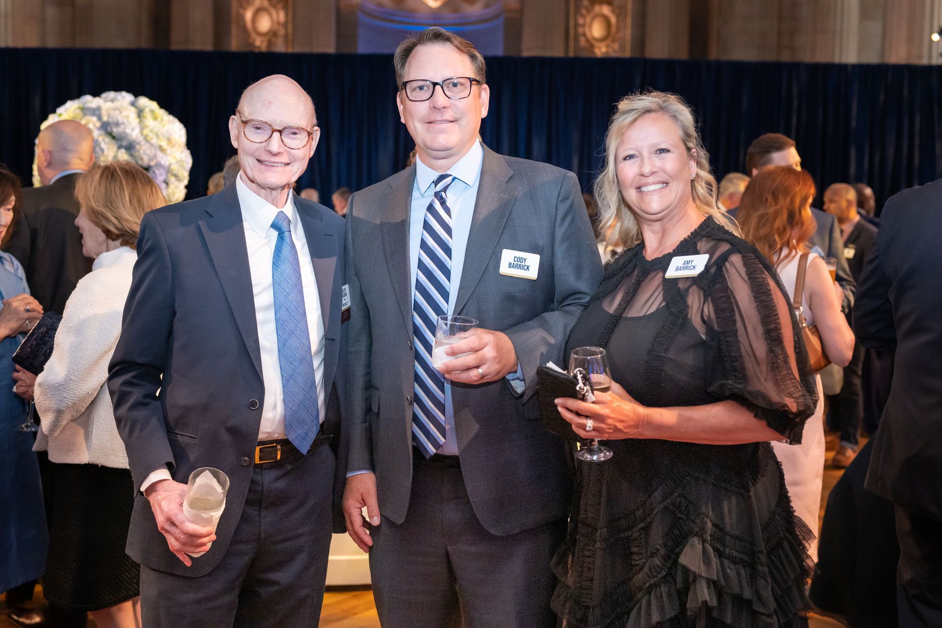 Three people in formal attire holding drinks at an indoor event, smiling for a photo