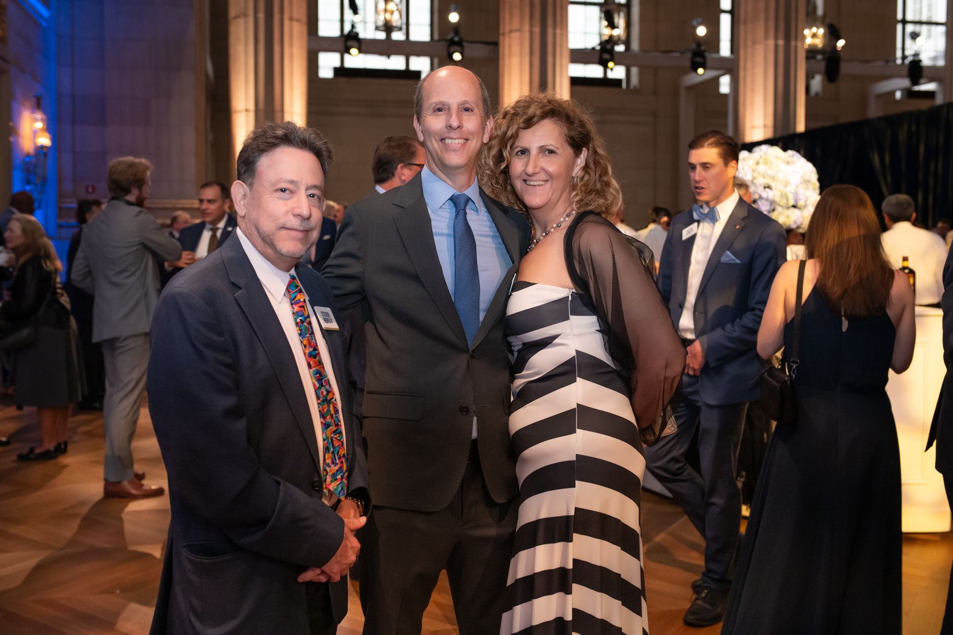 Three people posing at a formal indoor event, with guests and warm lighting in the background