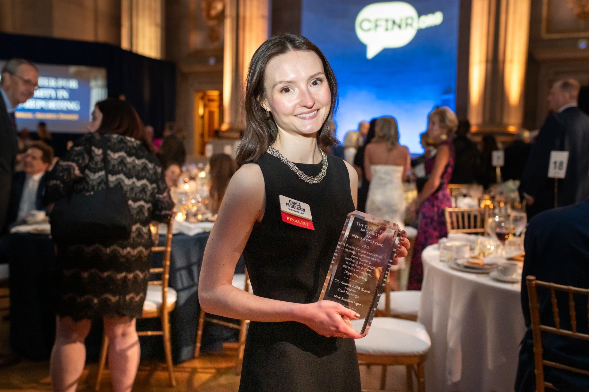Woman holding a plaque at a formal event, with guests and tables in the background.