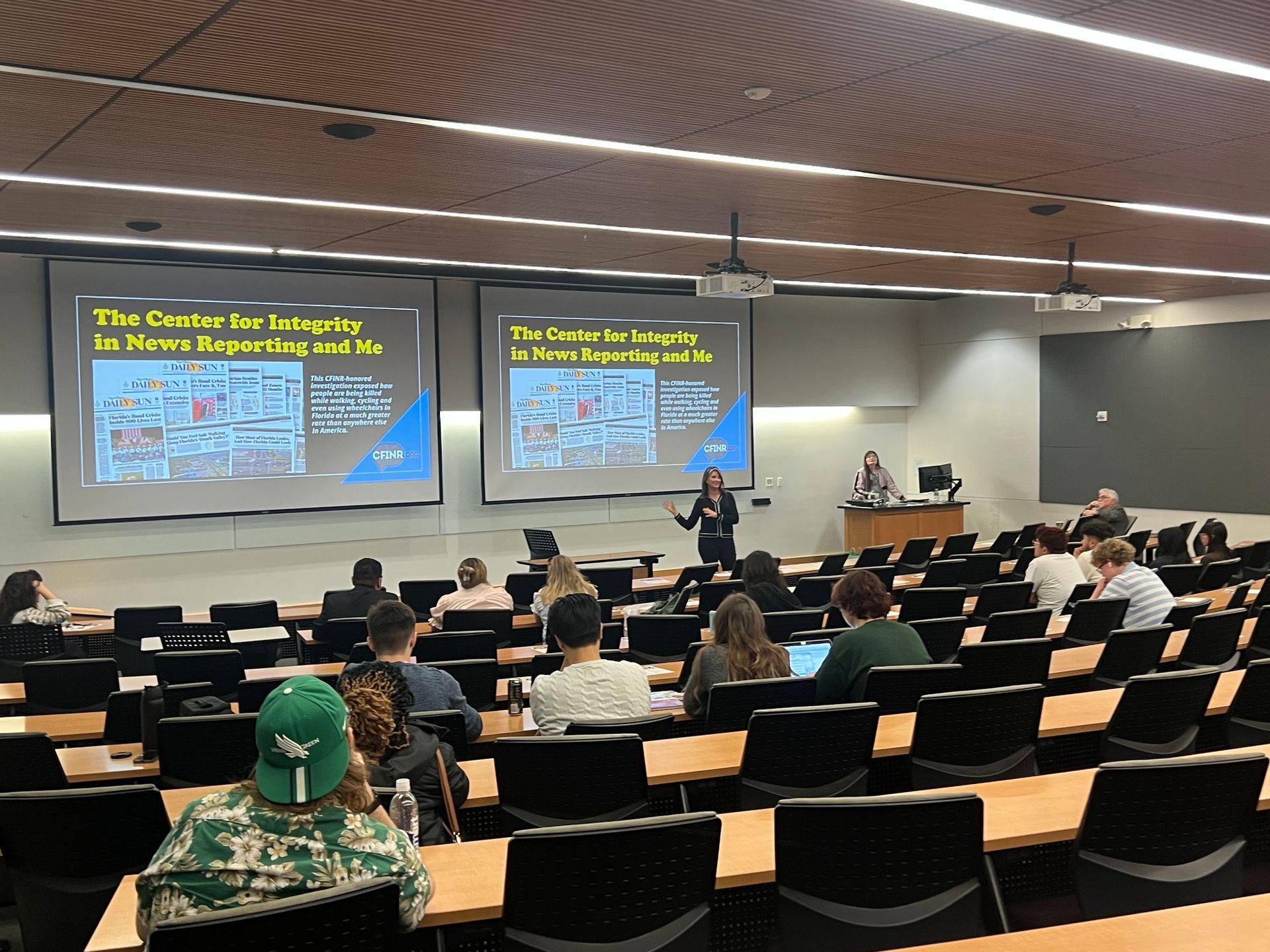 A lecture hall with a presenter and students; two screens display text.