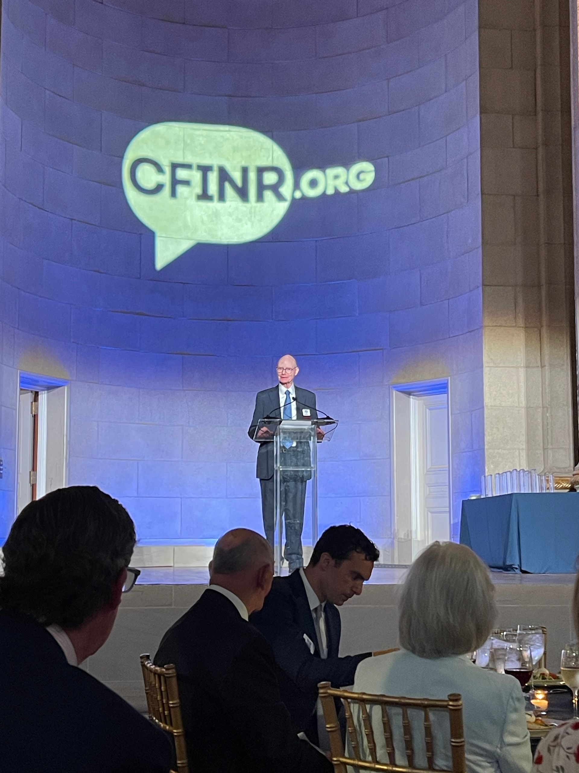 Speaker at podium on a stage with CFINR.org projected behind, viewed from a formal dinner audience.