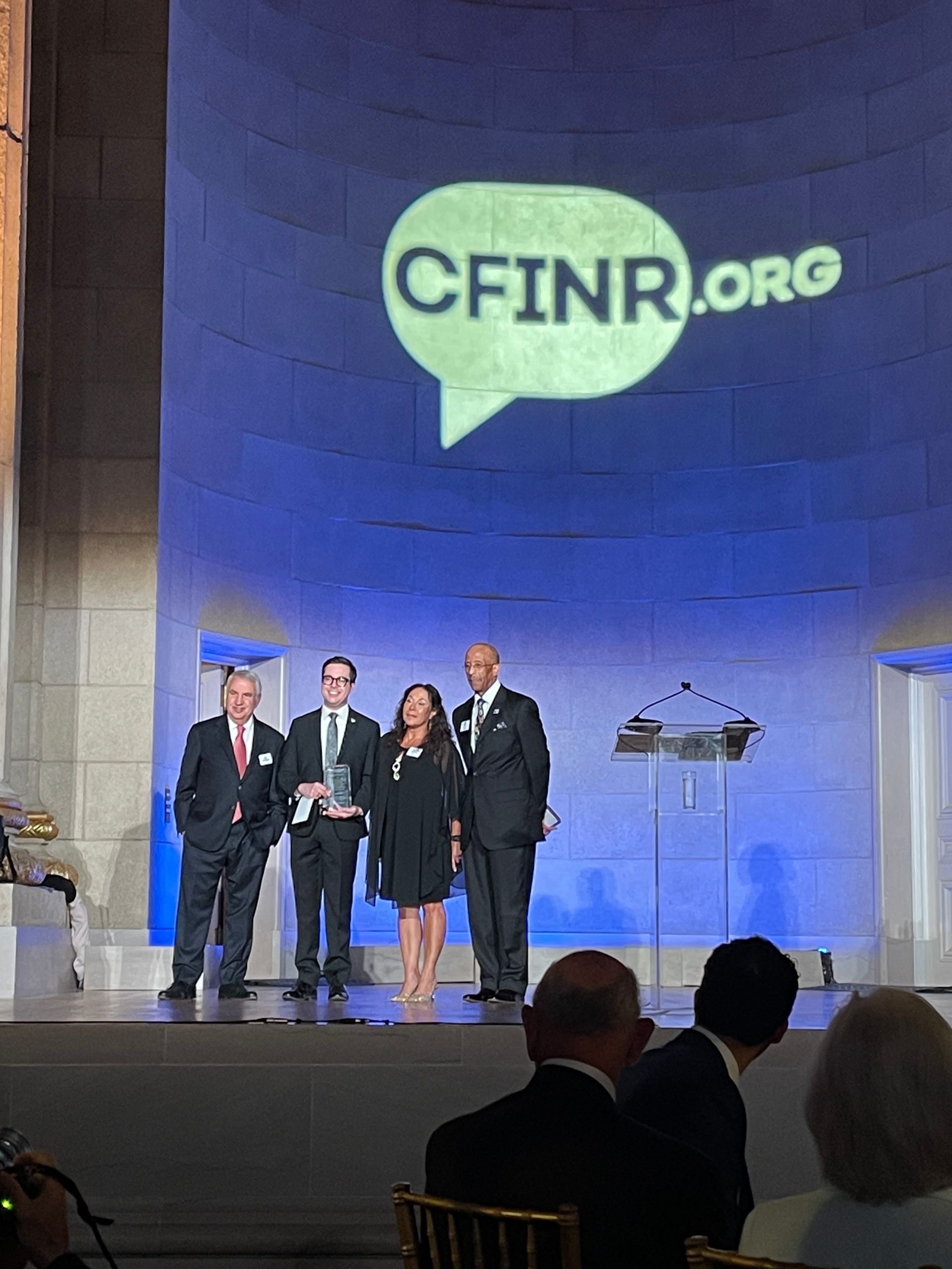 Four people on stage under a large CFINR.org logo, with an audience in the foreground.