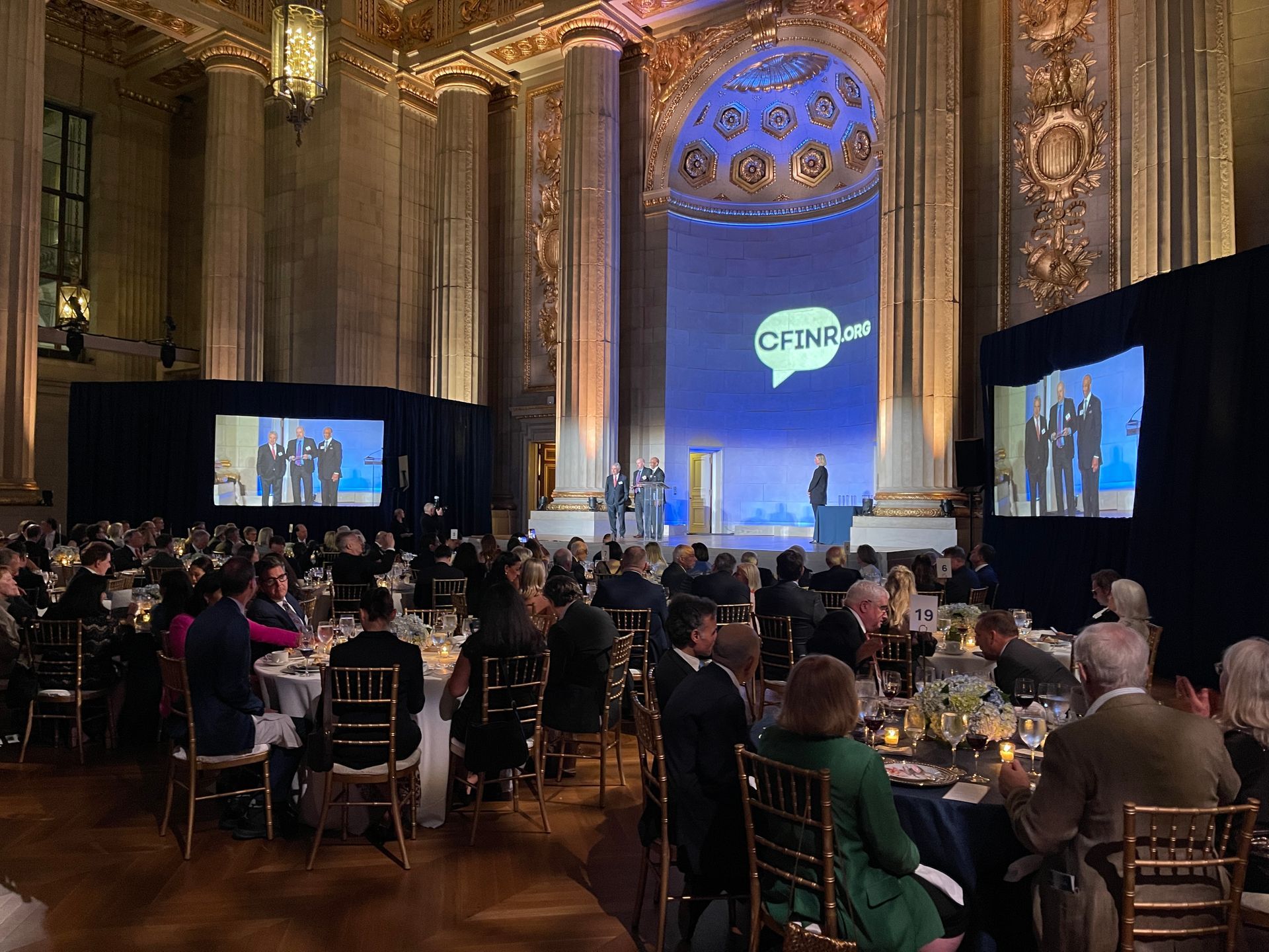 Banquet hall audience watching a stage presentation with large screens and a blue-lit backdrop