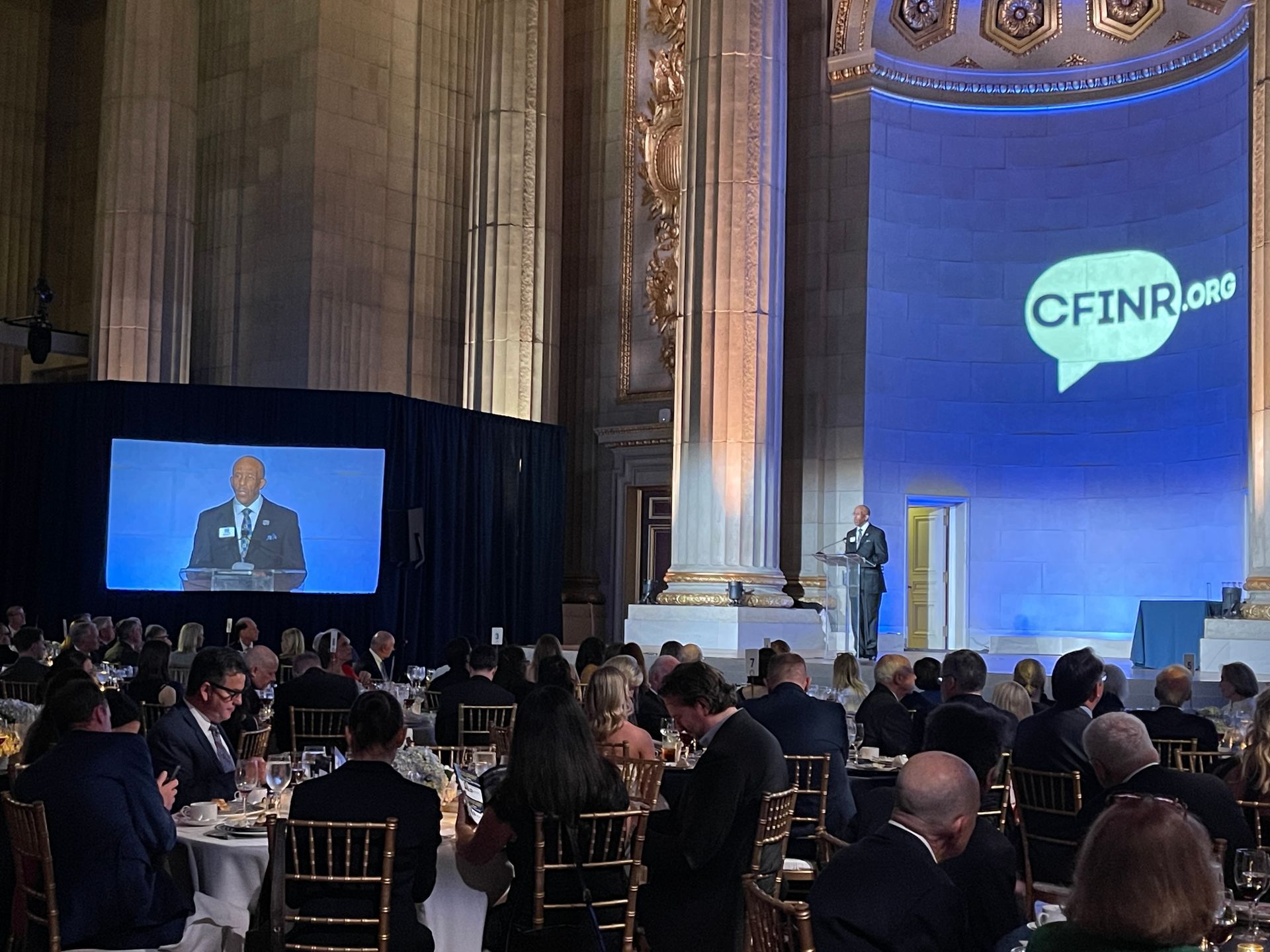 Audience watches a speaker onstage at a CFITN event in a grand hall with a large screen and blue-lit backdrop