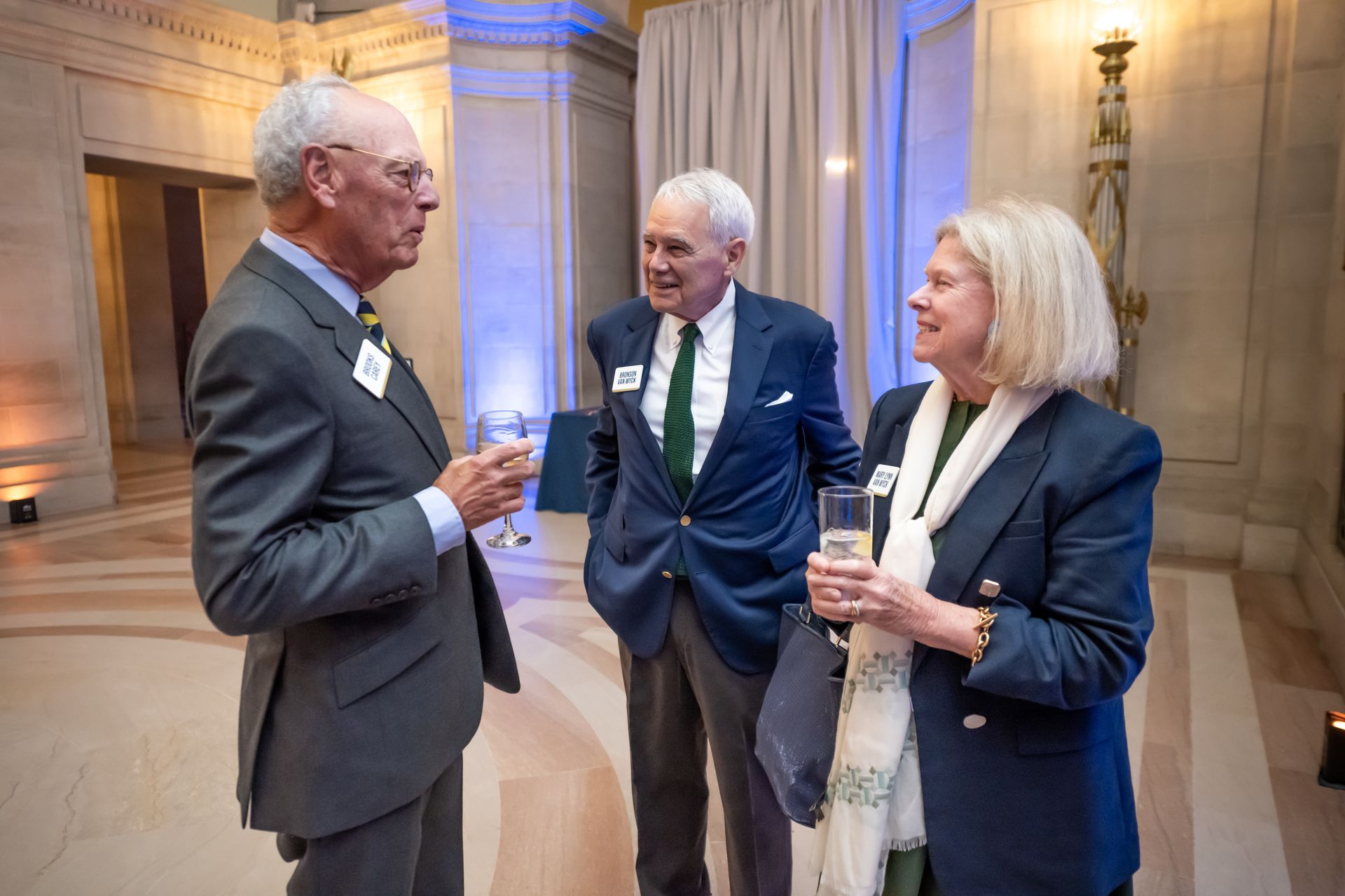 Three people in suits conversing in a grand hall, holding drinks and smiling.