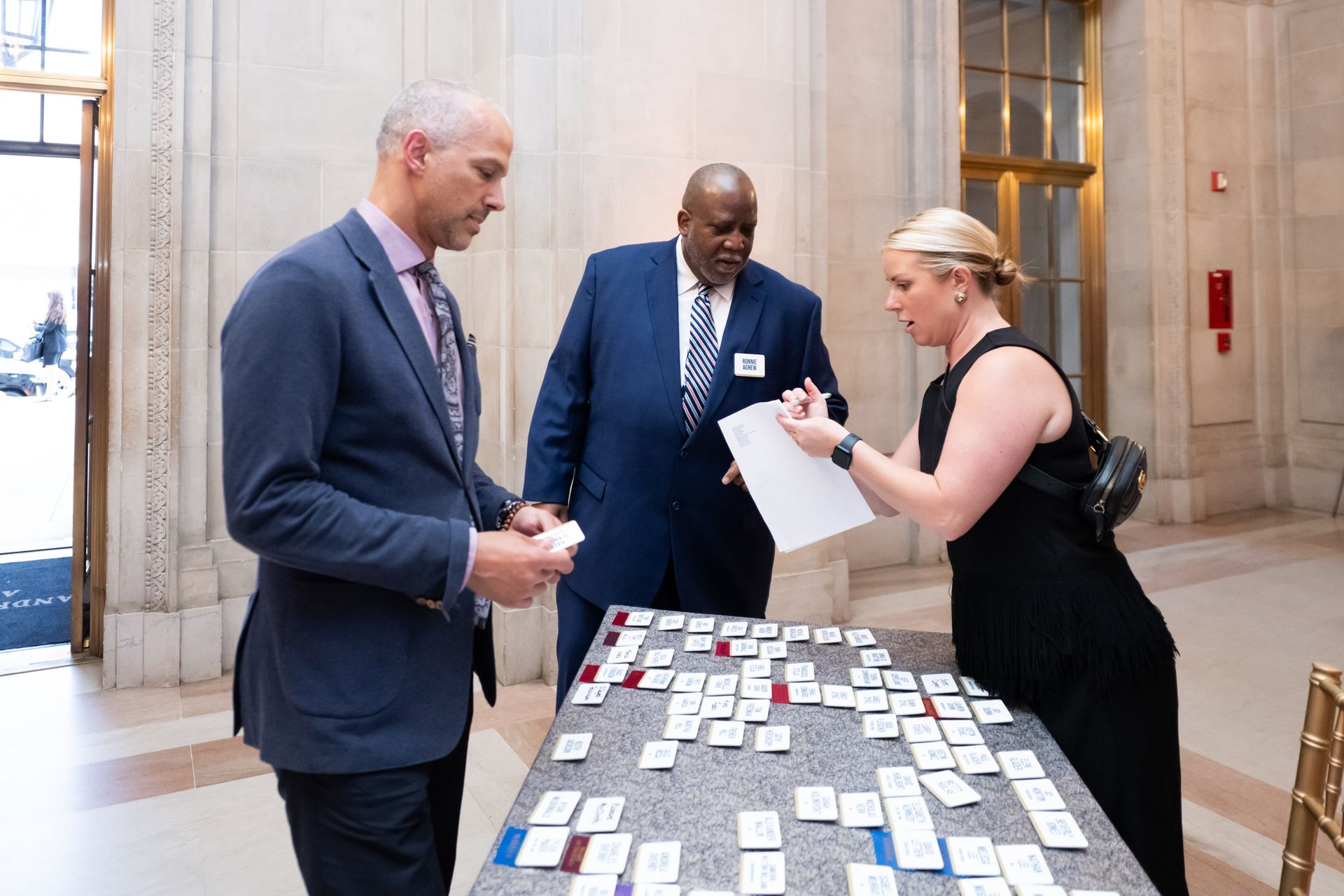 Three people stand beside a table covered with name tags, talking in a bright lobby.