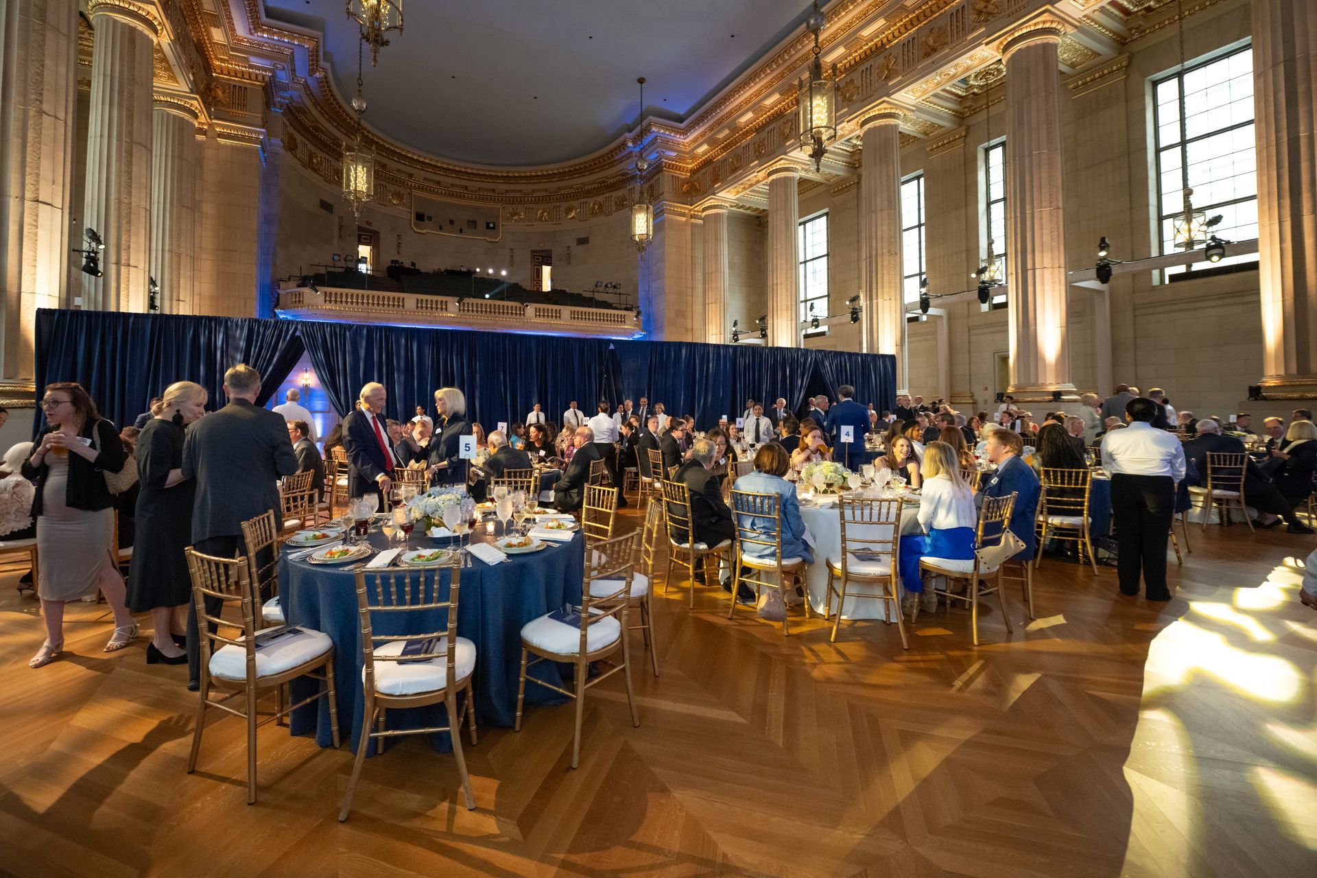 Formal banquet in a grand hall with round tables, seated guests, and blue drapes along the back wall