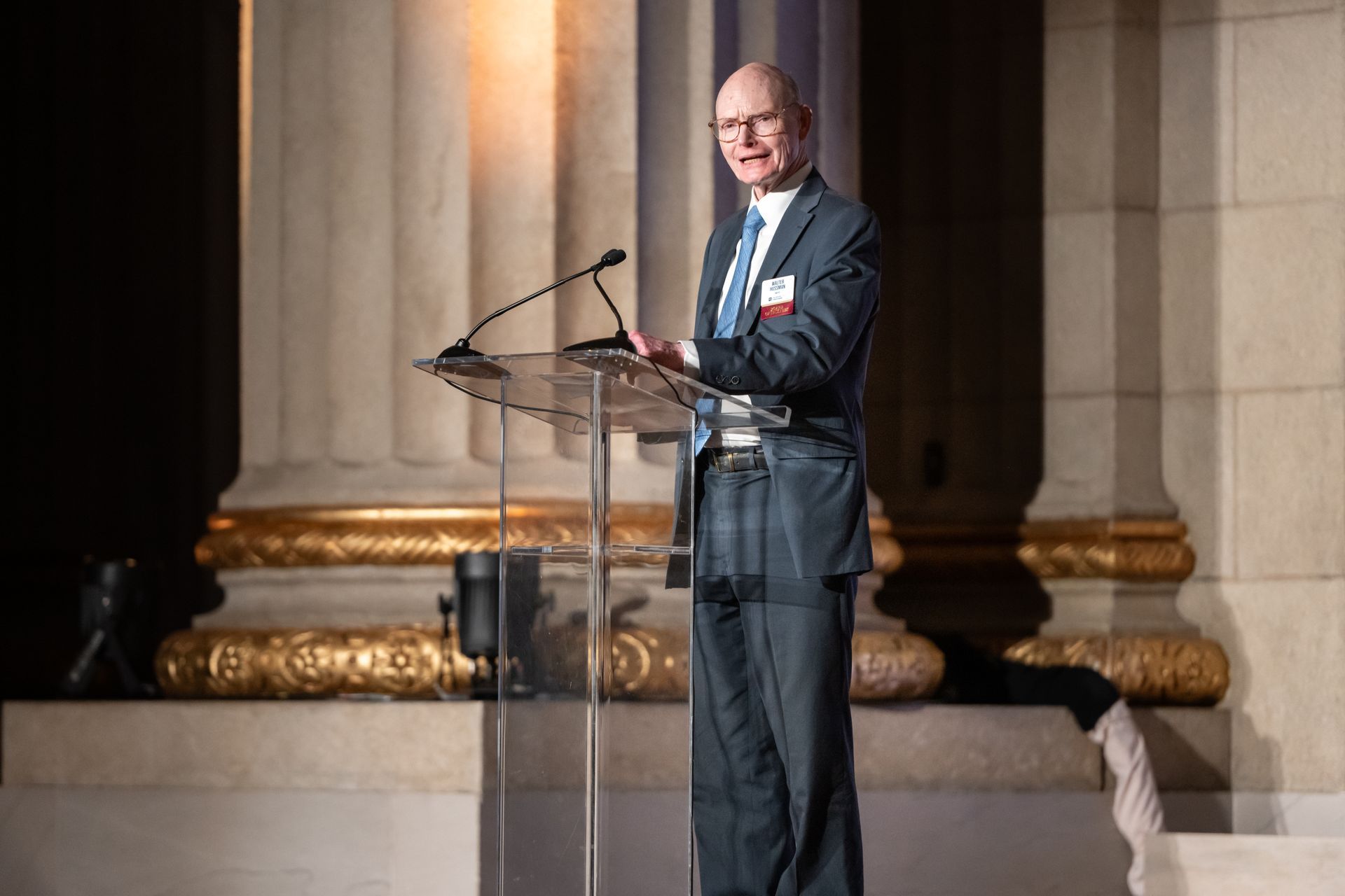 Man speaking at a podium in a formal hall with tall columns and warm lighting
