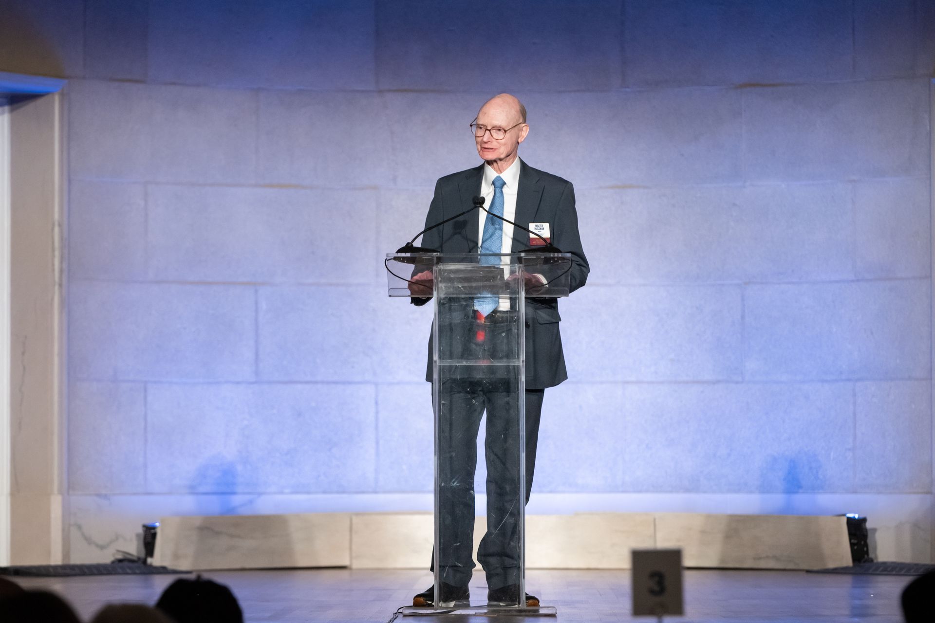 Man speaking at a podium on a stage with blue lighting