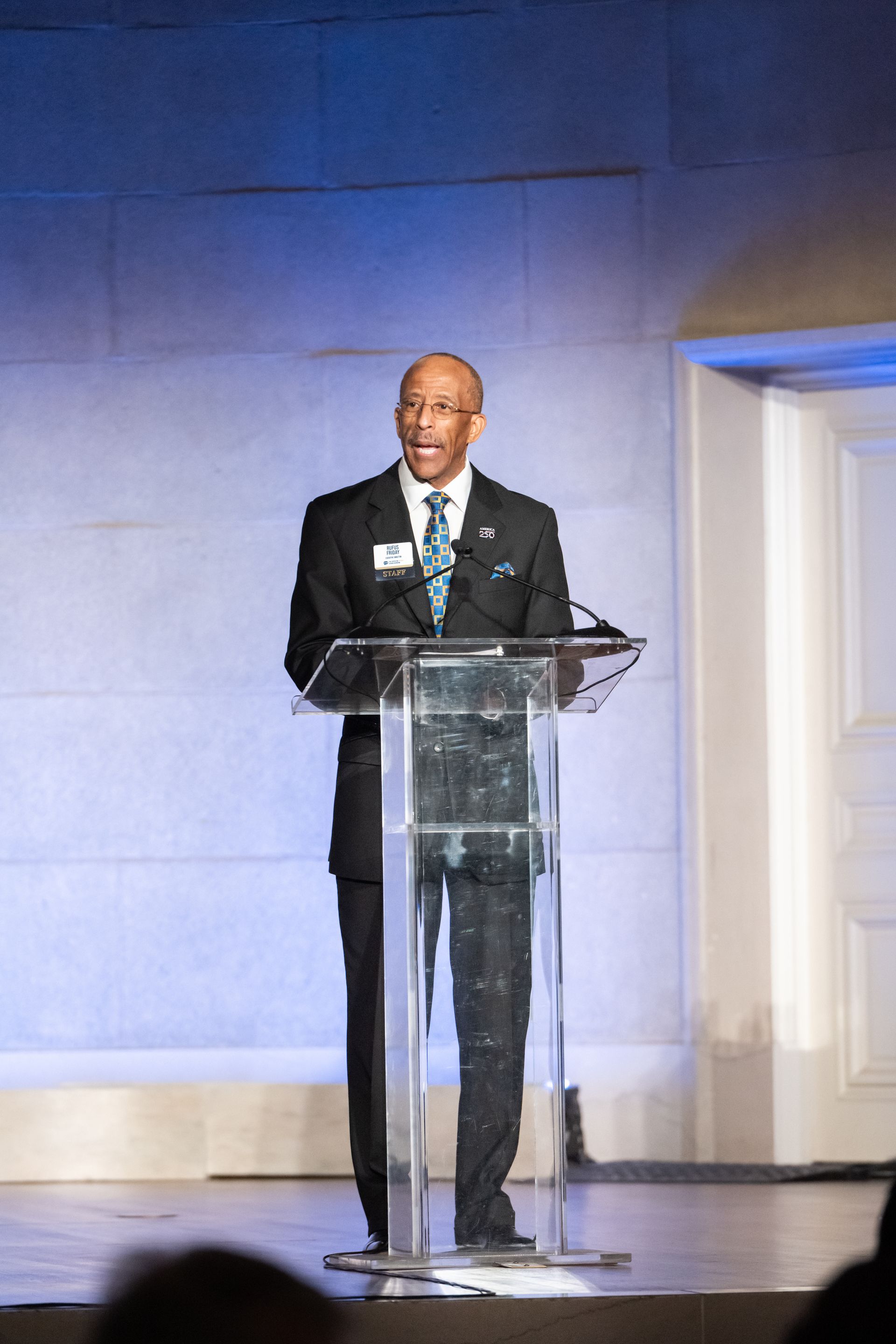 Speaker at a clear podium on a lit stage with blue backdrop and doorway.