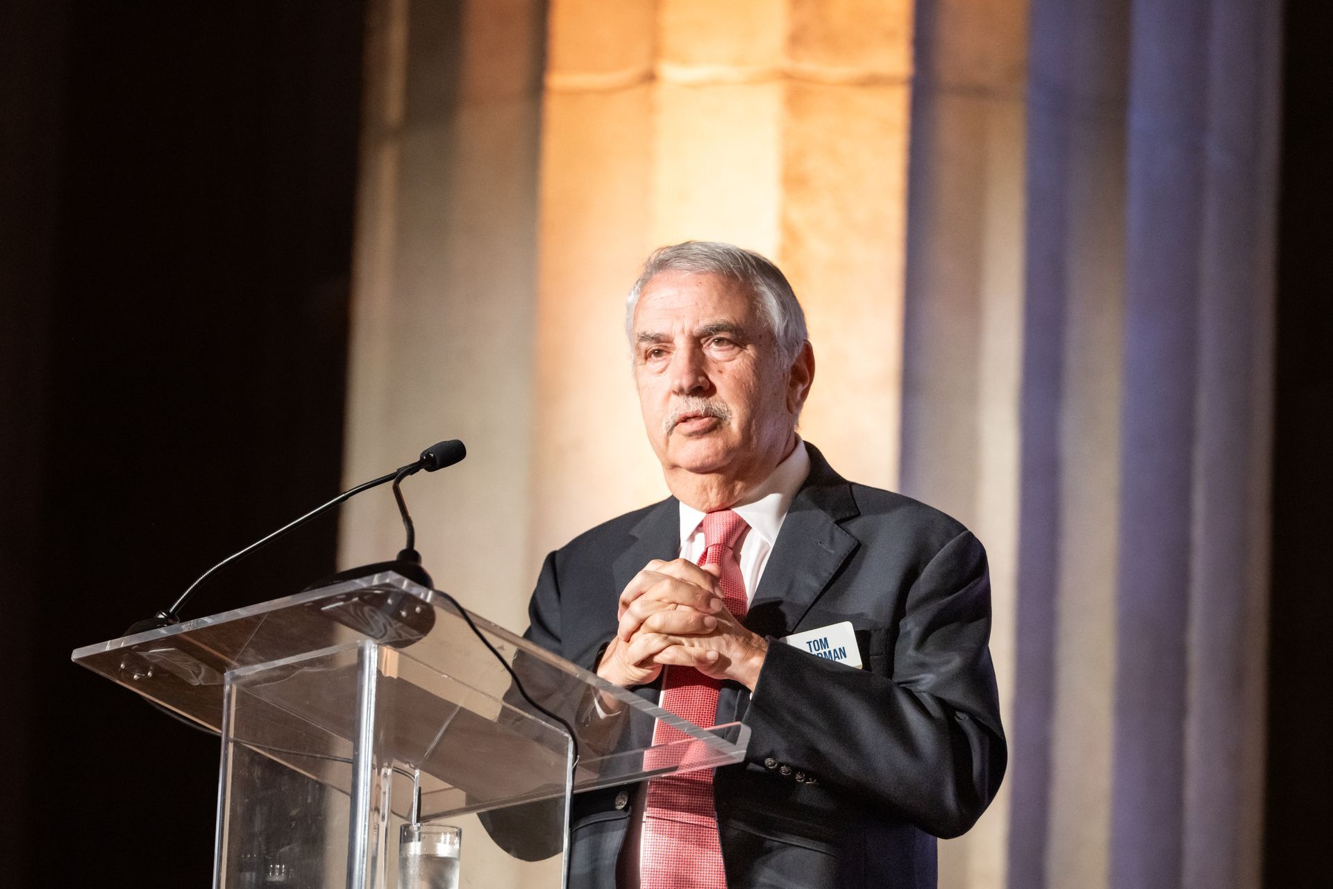 Man speaking at a podium with a microphone, wearing a dark suit and red tie, against a softly lit stage background