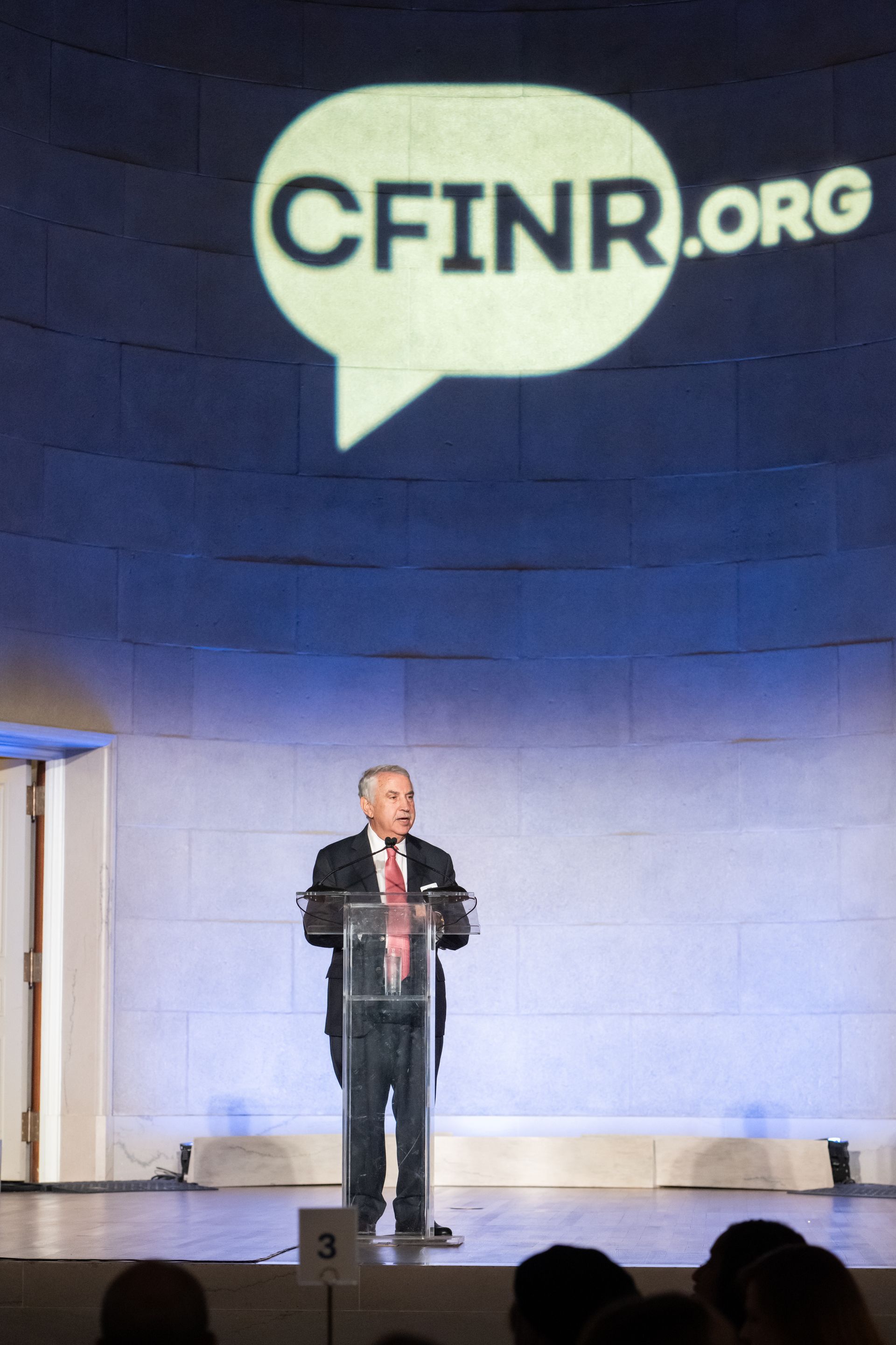 Speaker at podium on stage beneath large CFINR.org logo projected on a blue wall.
