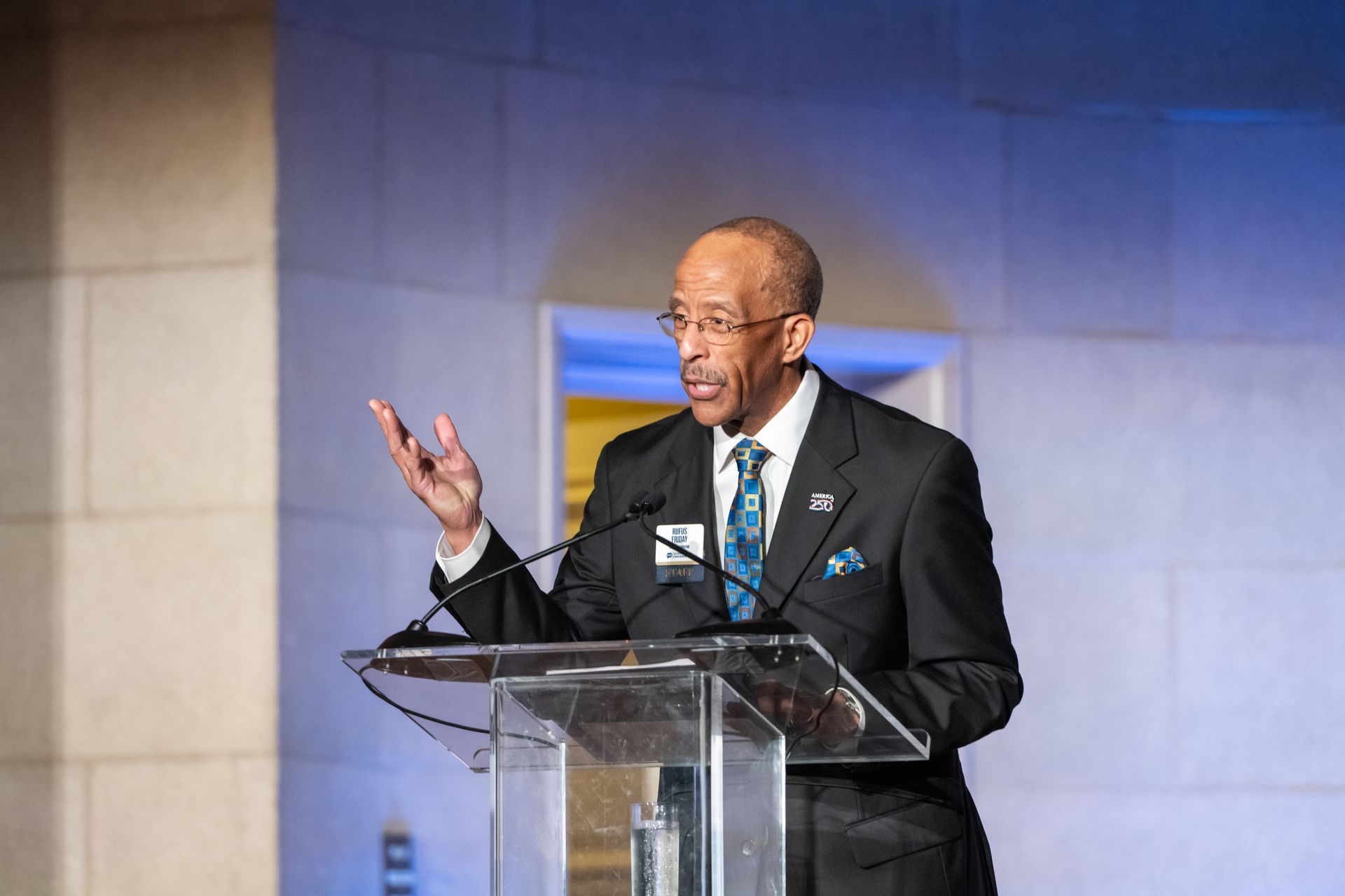 Man speaking at a podium with microphones, gesturing during a formal event in a lit hall