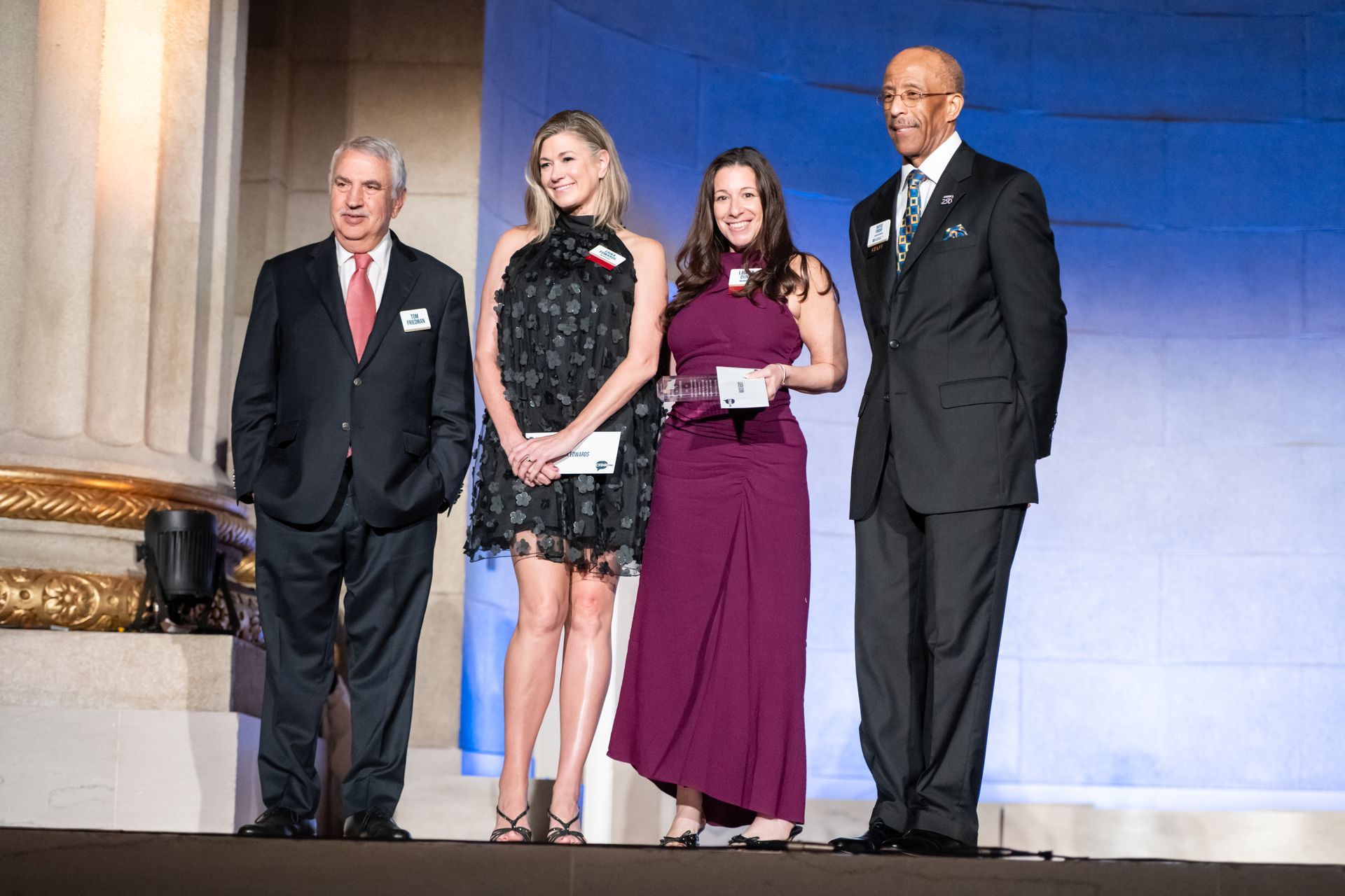 Four people stand on a stage in formal attire, one holding a plaque, at an awards ceremony.