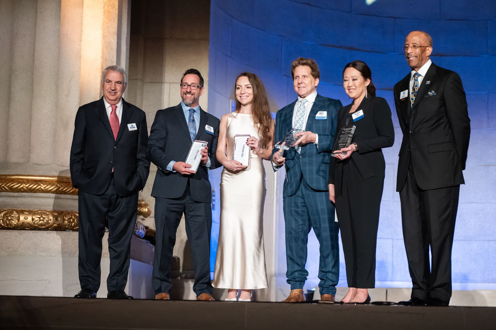 Six people in formal wear pose holding awards on a stage with ornate columns.