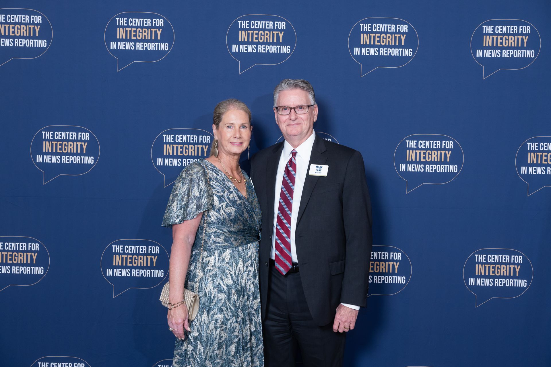 Two people posing in formal attire before a blue event backdrop with logos