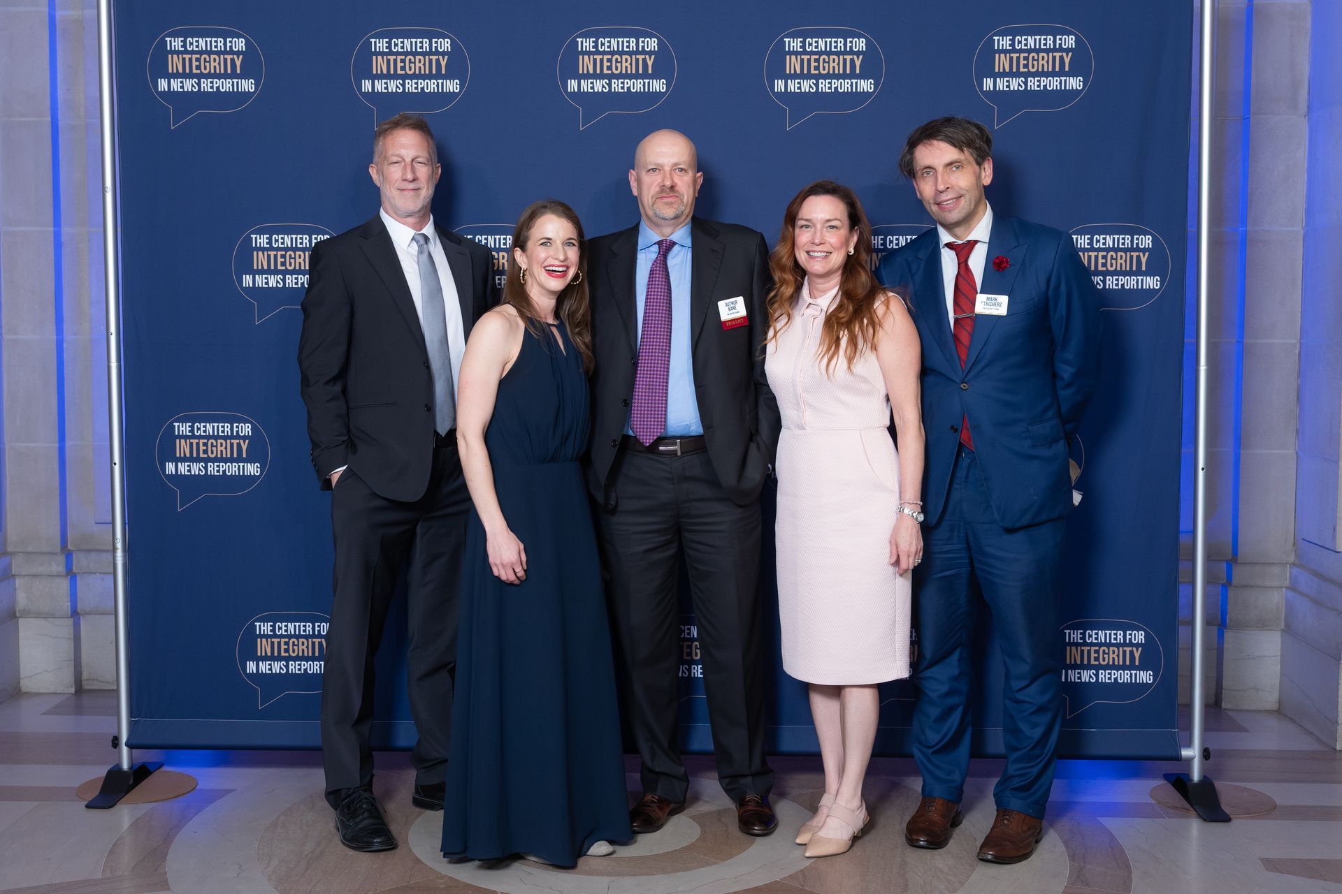 Five people posing on a blue step-and-repeat event backdrop, dressed in formal attire.