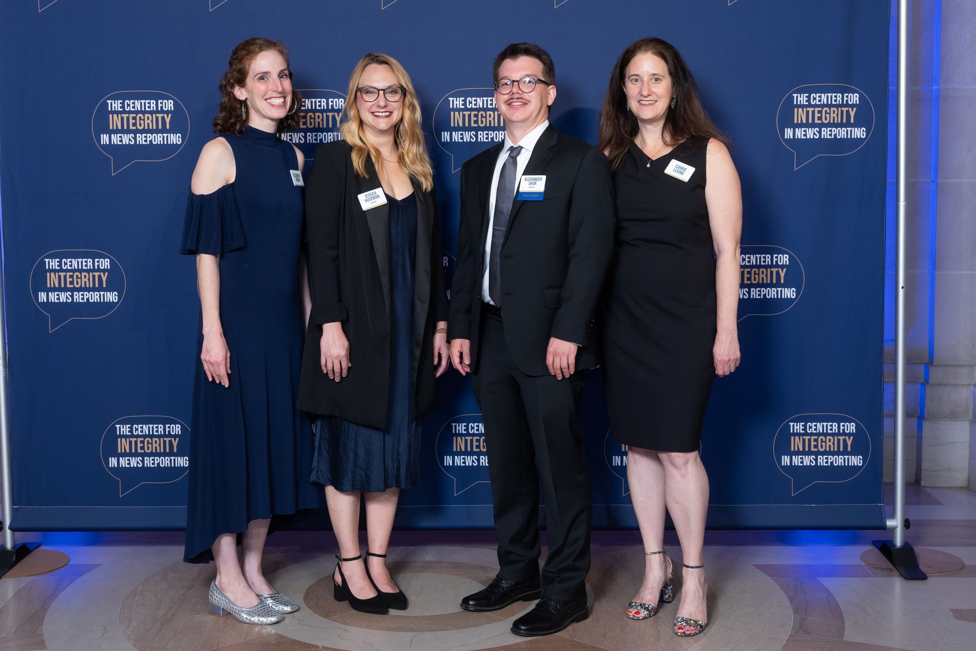 Four people posing in formal attire in front of a blue event backdrop with logos.
