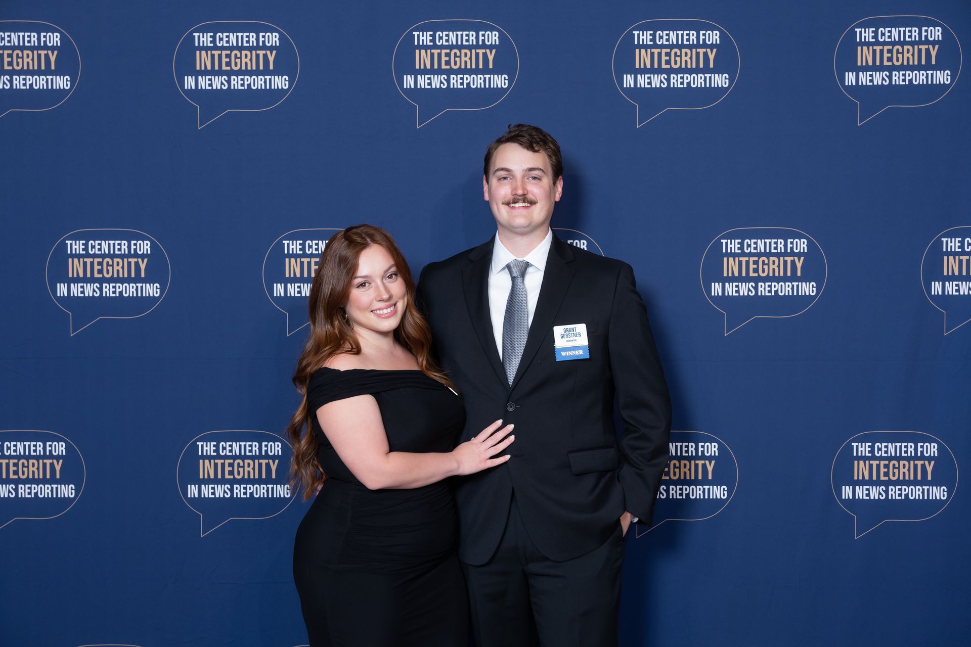 Two people posing together at an awards event in front of a blue step-and-repeat backdrop