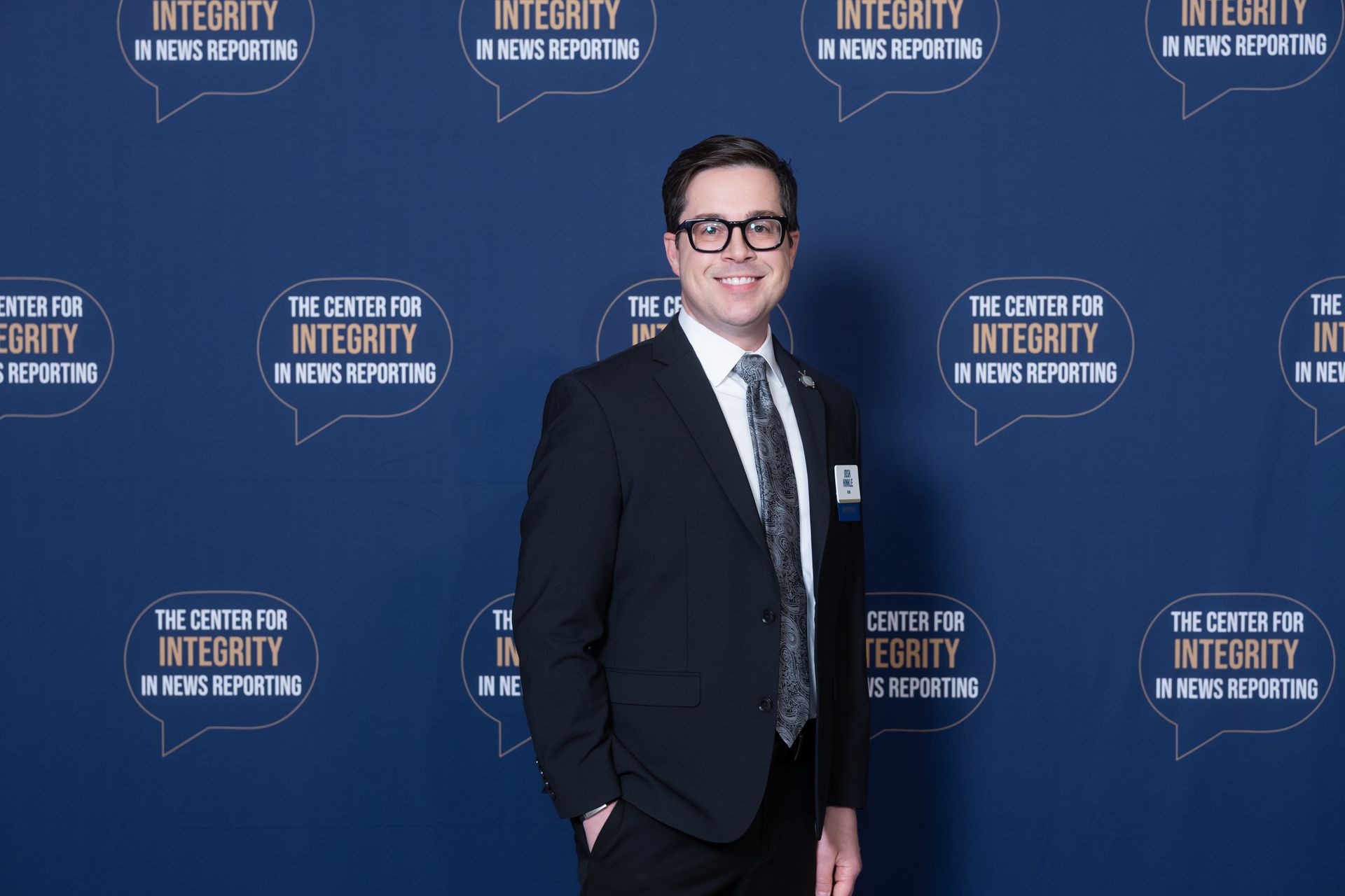 Man in a dark suit and glasses stands smiling before a blue Integrity Summit backdrop.