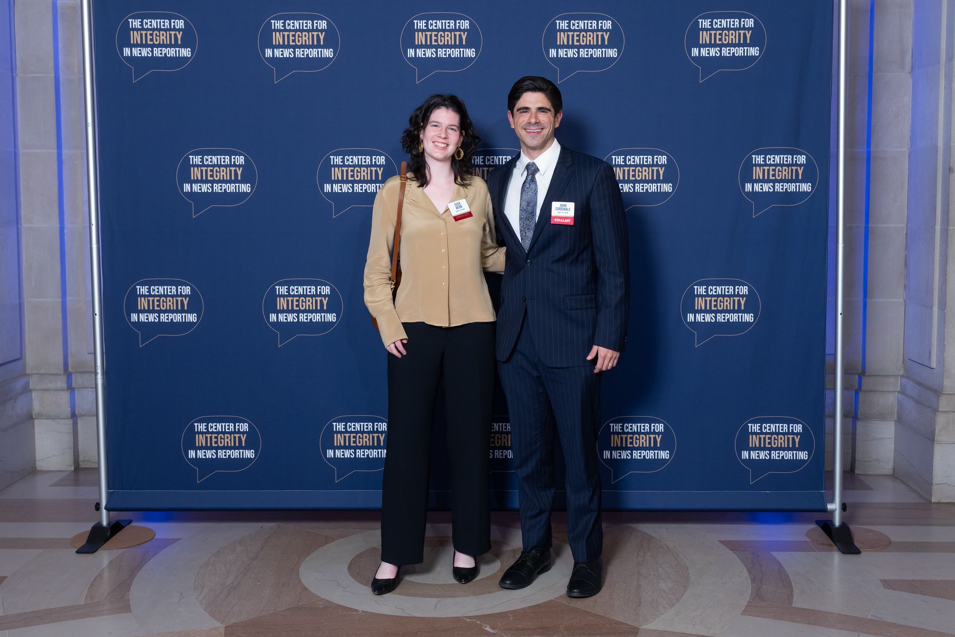 Two people posing in front of a blue event backdrop with logos, smiling and wearing name tags.