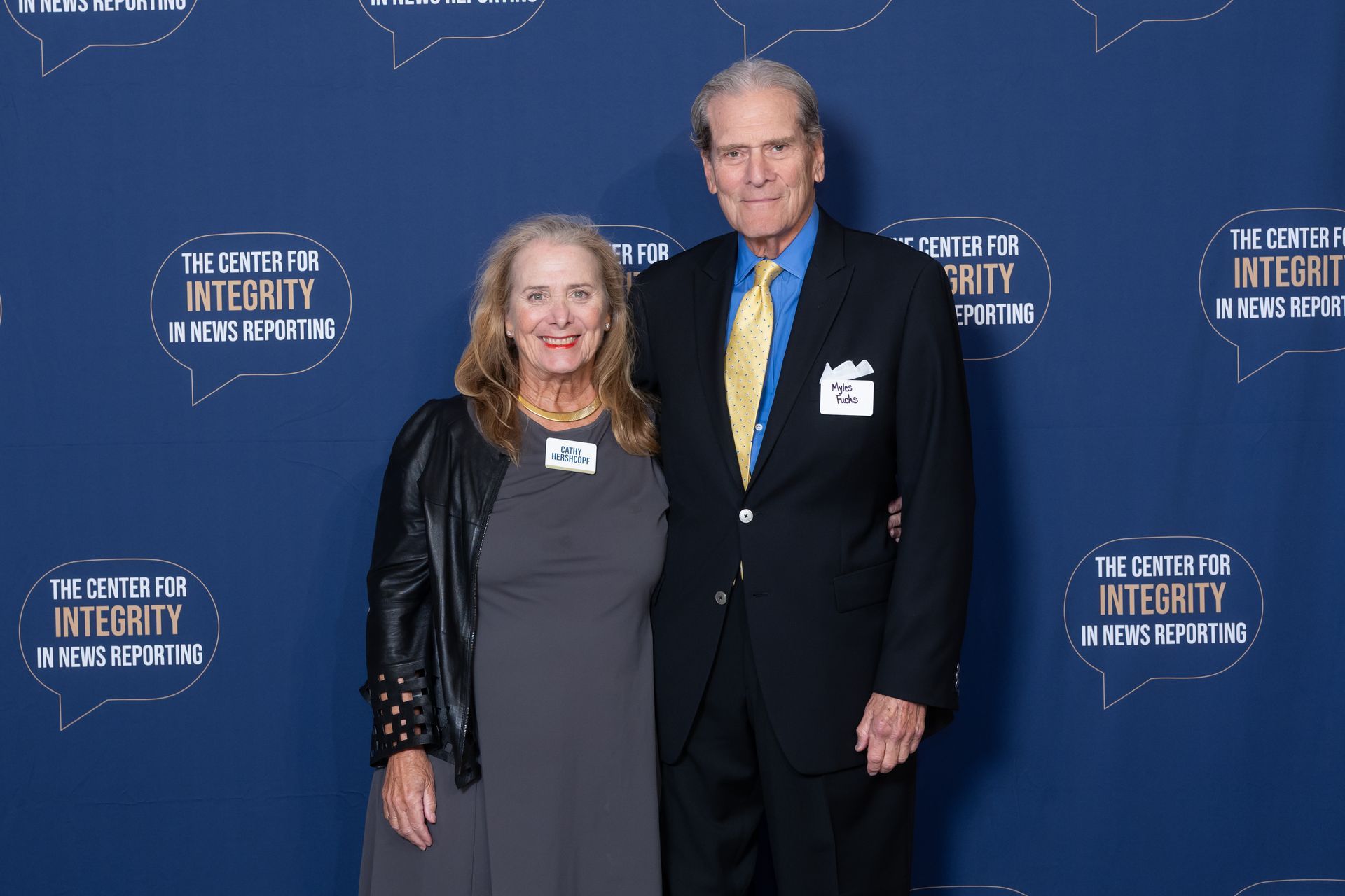 Two people posing on a blue event backdrop, both in dark formal attire.