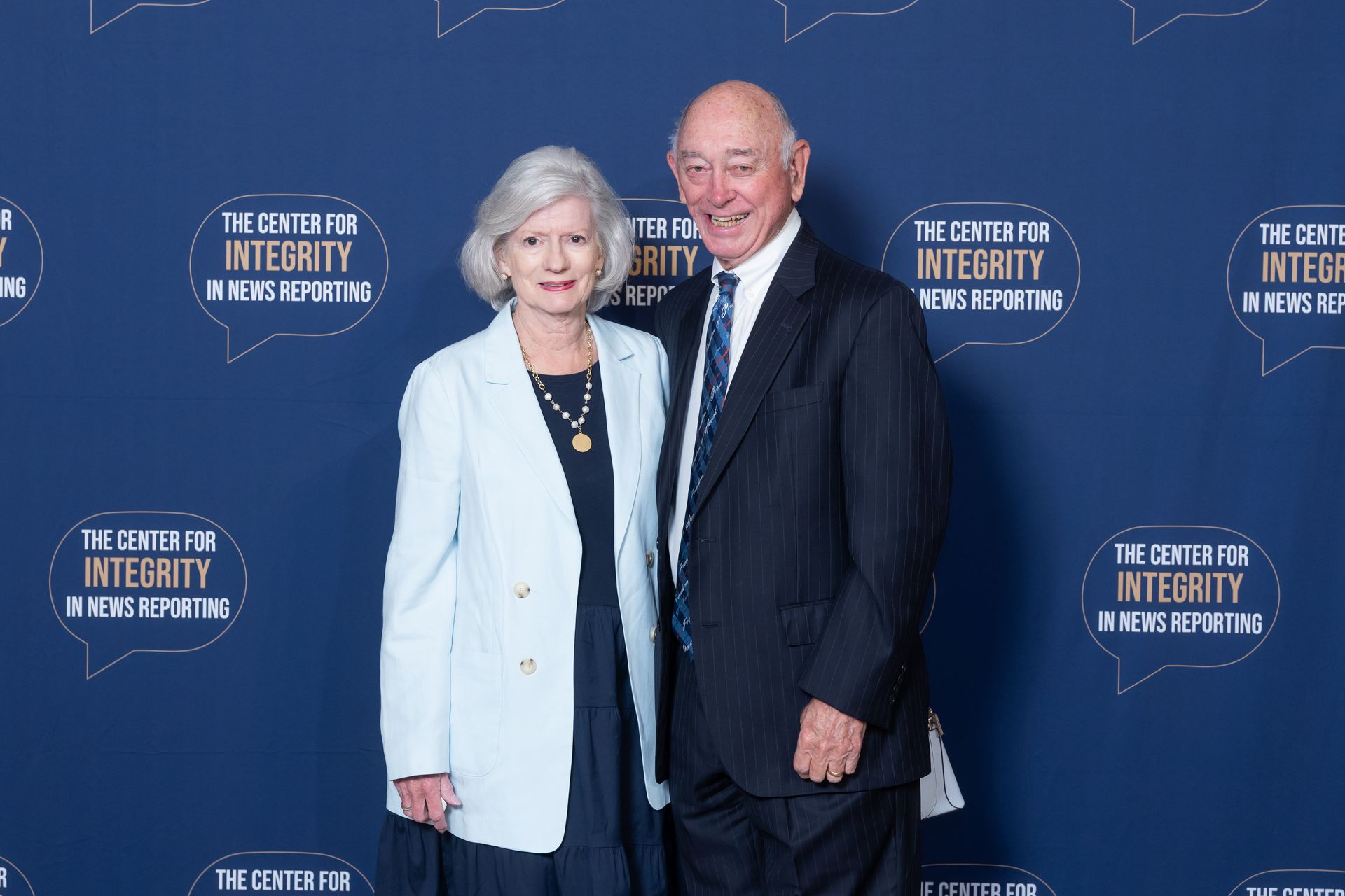 Two people posing at a blue-step-and-repeat event backdrop, both smiling and dressed in formal attire.
