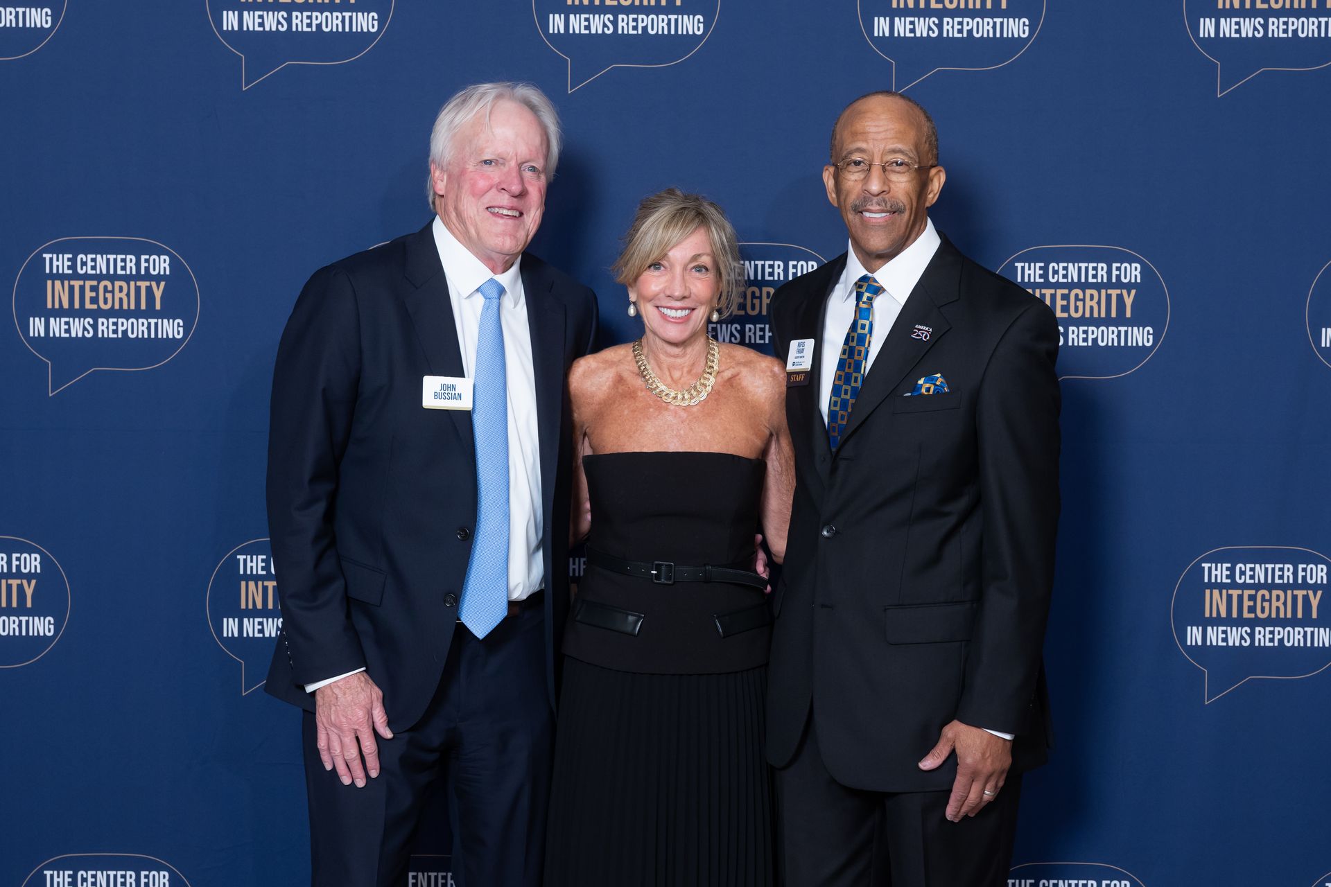 Three people posing at a blue step-and-repeat event backdrop, smiling in formal attire.