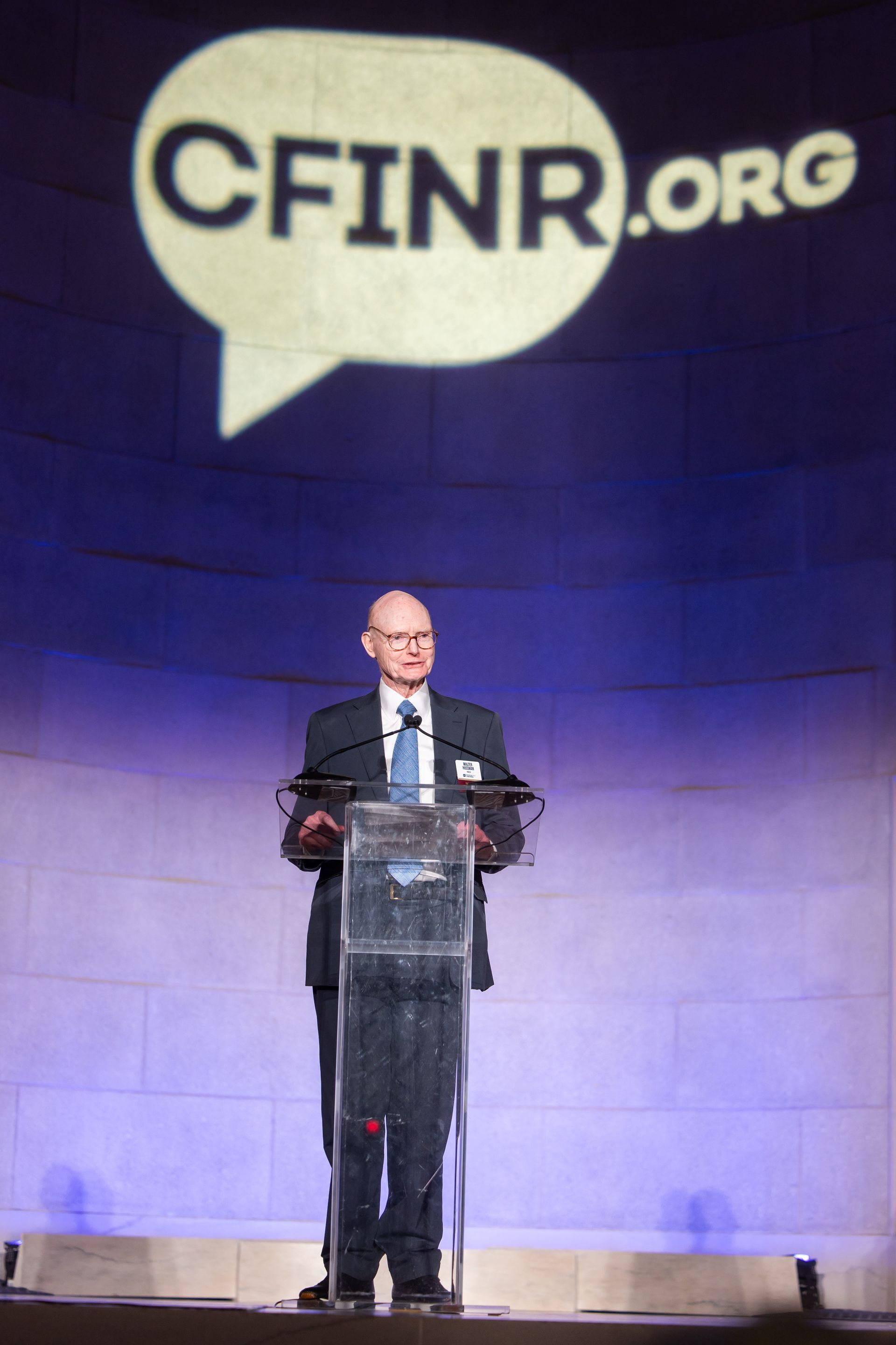 Speaker at a podium on stage beneath a large CFINR.org logo projection.