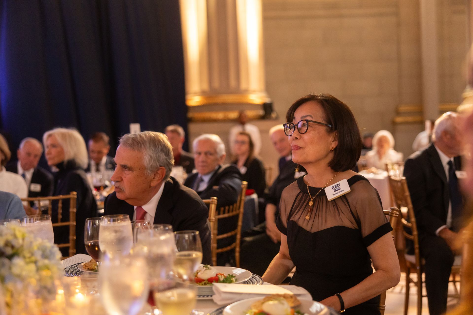 Formal banquet audience seated at round tables, listening in an elegant, warmly lit hall.