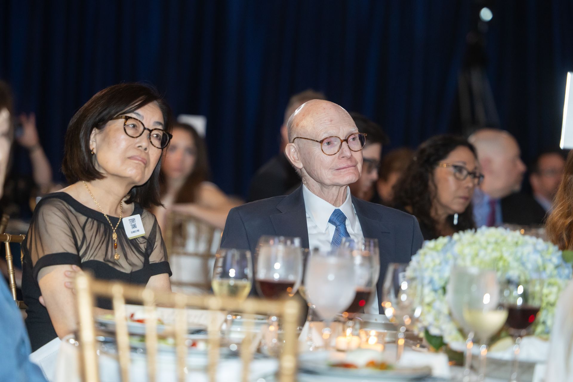 Attendees seated at a formal dinner table, listening during an event with wine glasses and floral centerpieces