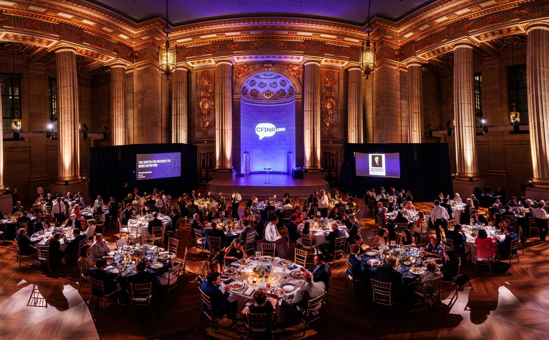 Elegant banquet hall with round tables, seated attendees, and a stage lit in purple and gold.