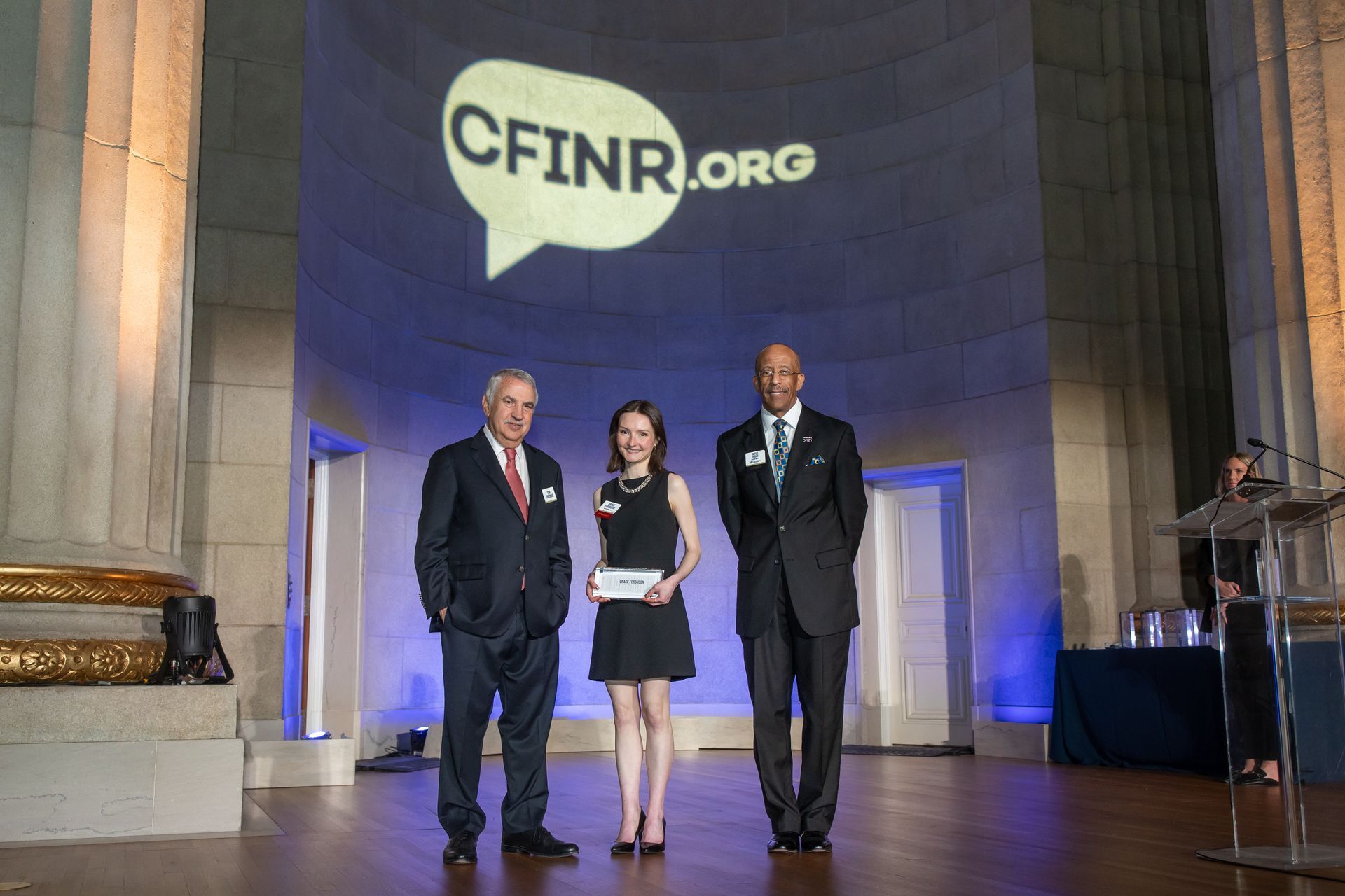Three people on stage at a CFINR.org award ceremony, one holding a plaque under blue lighting.