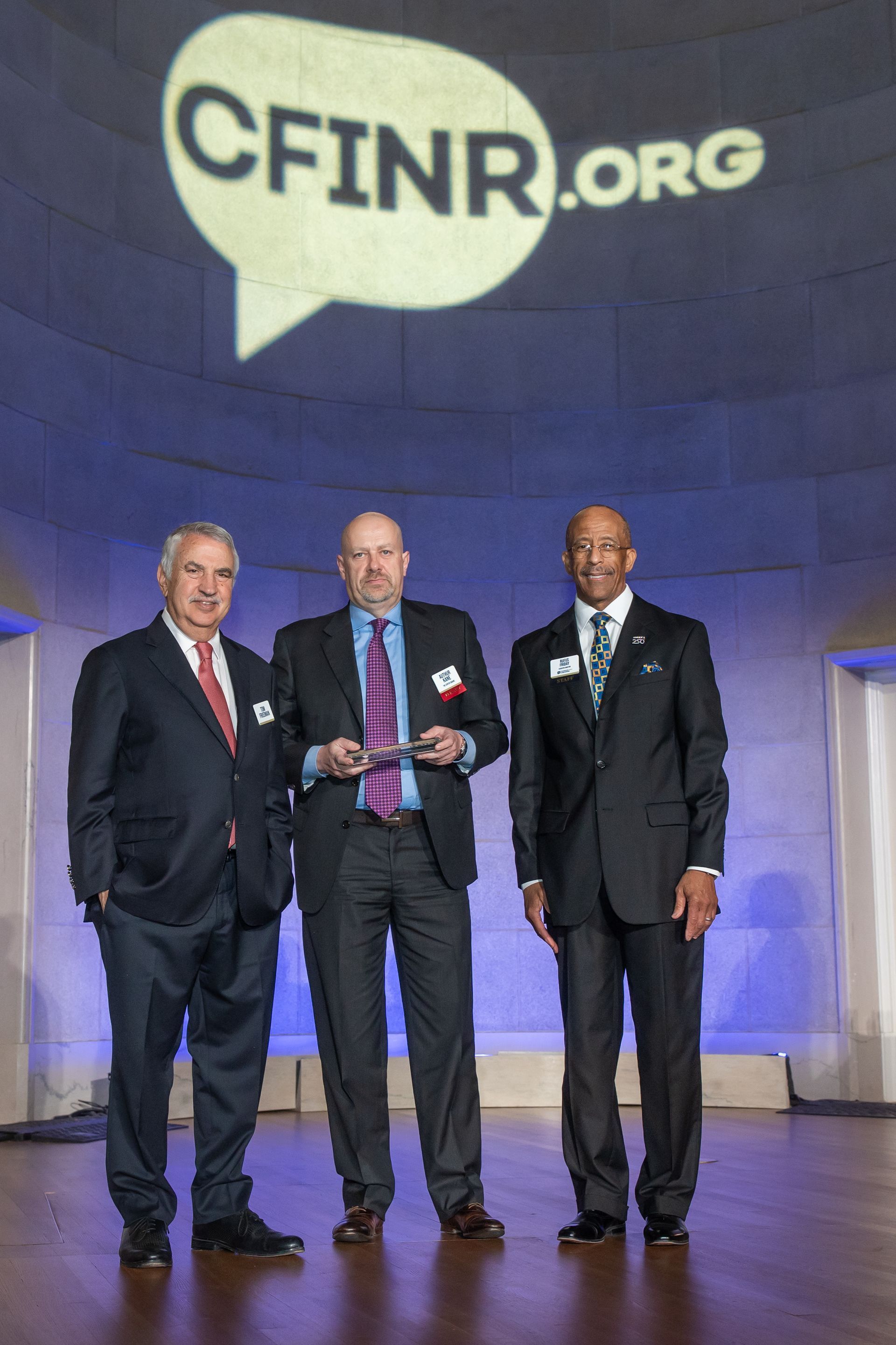 Three men in suits standing onstage beneath the CFINR.org logo, one holding an award