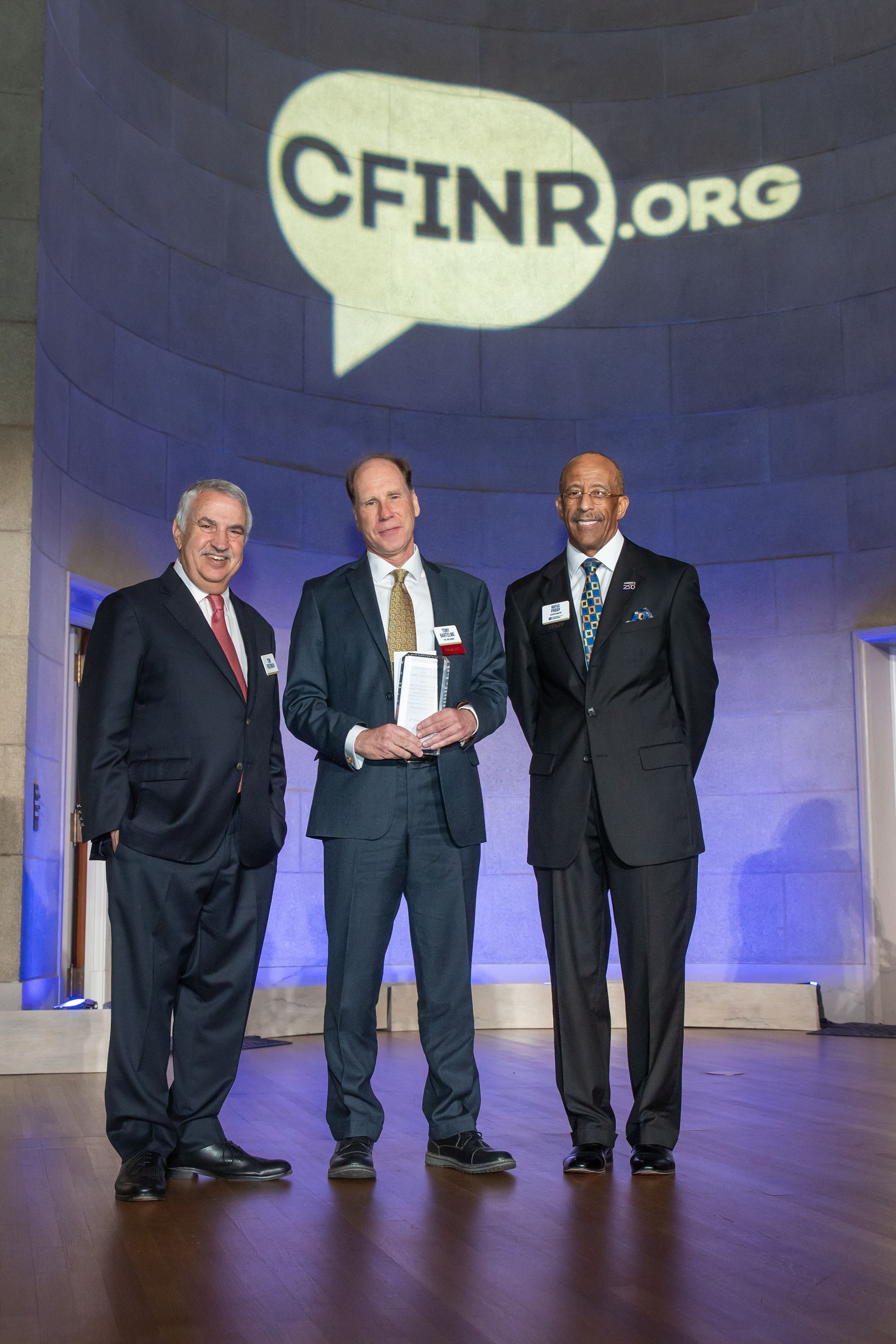 Three men in suits standing onstage before a large CFINR.org backdrop